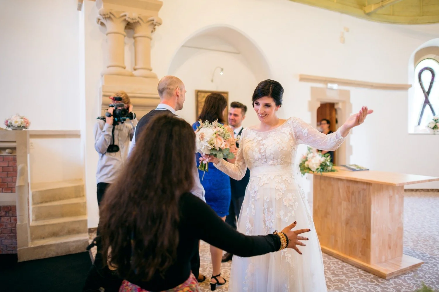 Nevěsta s kyticí otevírá náruč ženě přicházející uličkou, zatímco v pozadí stojí další lidé a fotograf.