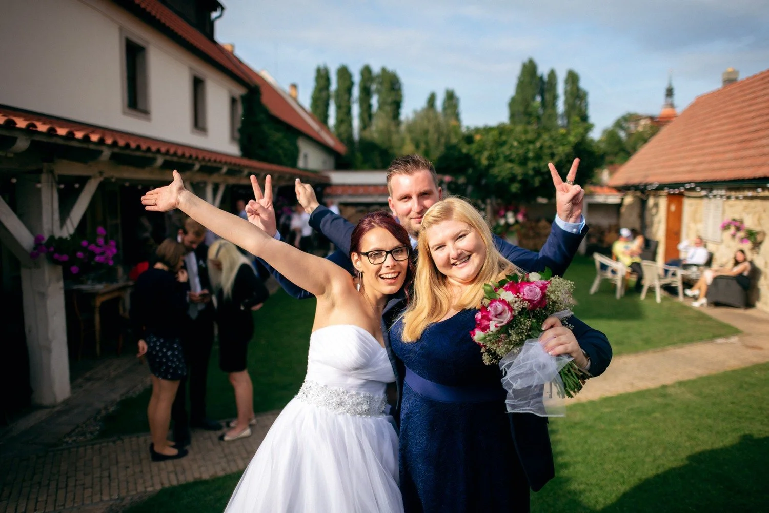 Žena v bílých šatech a blondýna s kyticí radostně zvedají ruce na zahradě.