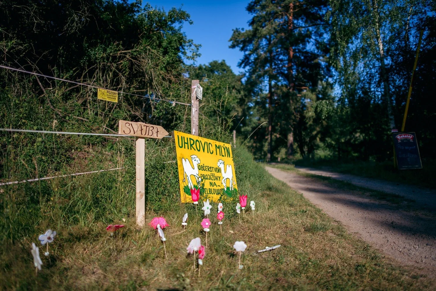 U prašné cesty stojí dřevěný rozcestník ozdobený barevnými papírovými květy.