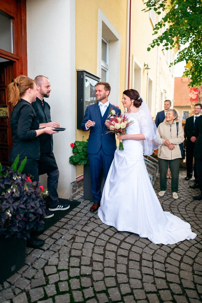 Dvojice ve svatebním oblečení vychází na dlážděný chodník mezi lidmi stojícími u vchodu.