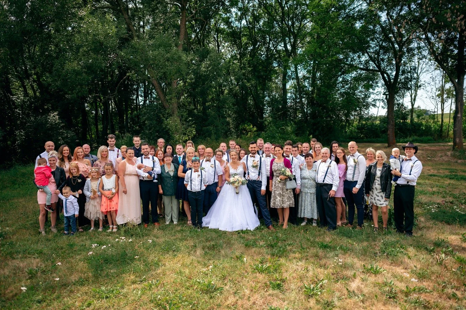 Nevěsta se ženichem stojí uprostřed velké skupiny hostů na kraji lesa.