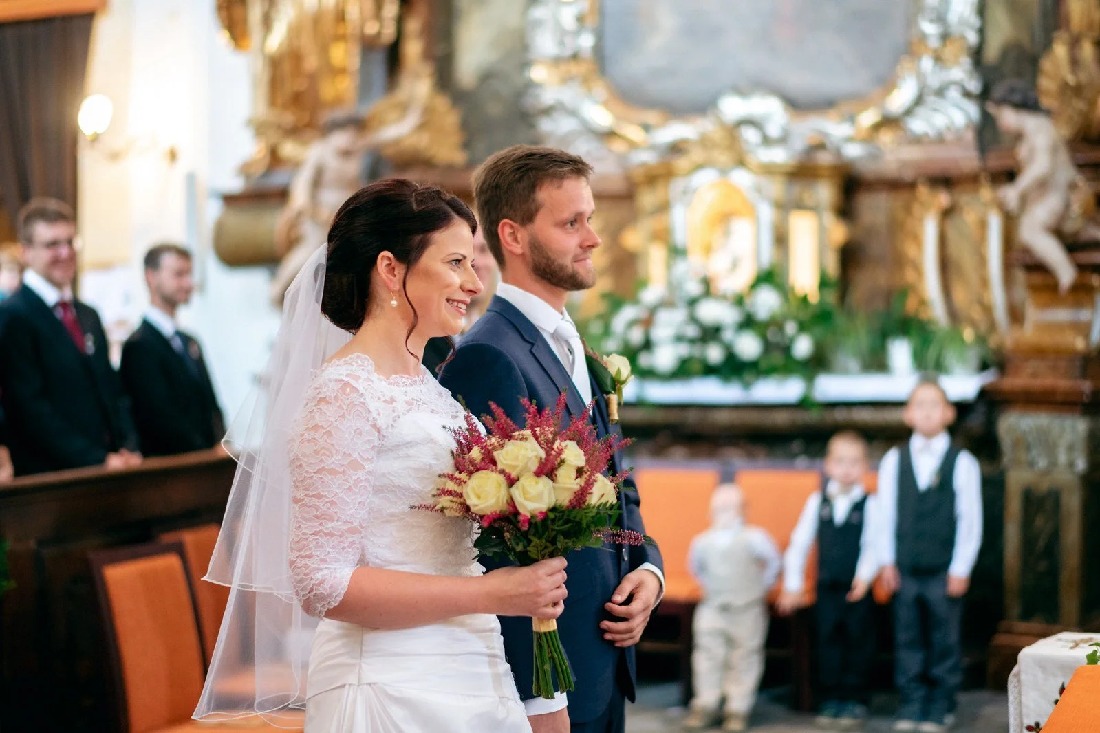 Dvojice ve svatebním oblečení stojí s kyticí v popředí před květinově zdobeným oltářem.