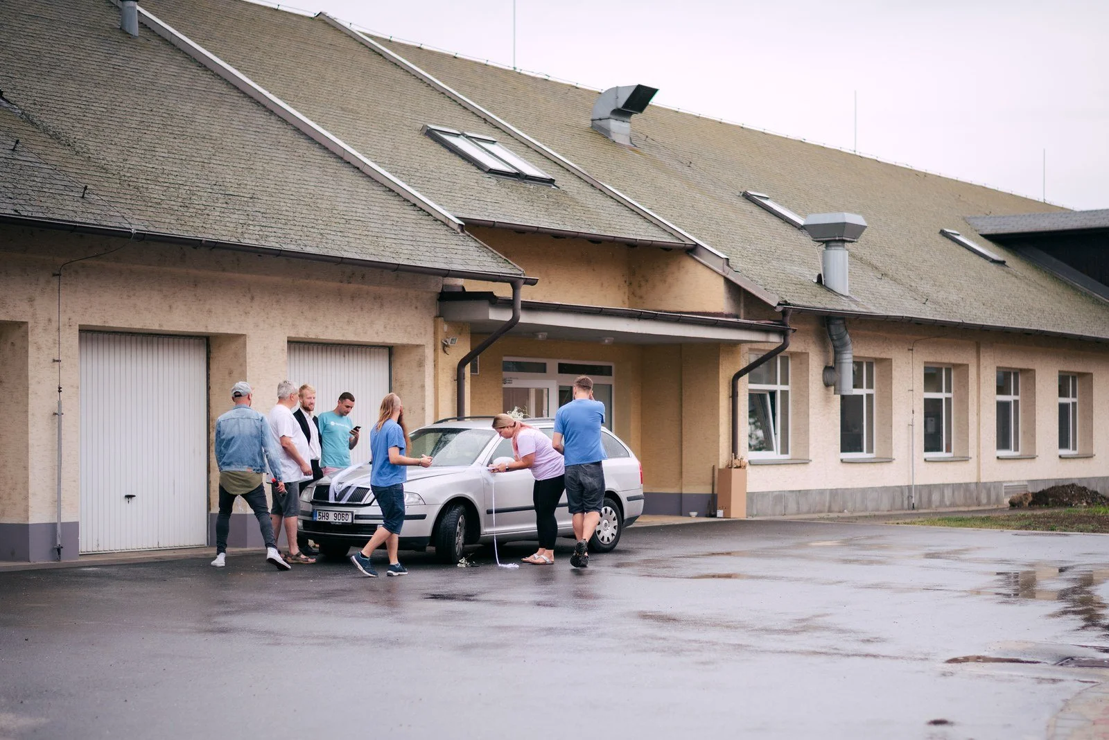 Před dlouhou světlou budovou stojí malá skupina lidí zachycená v širokém venkovním záběru.