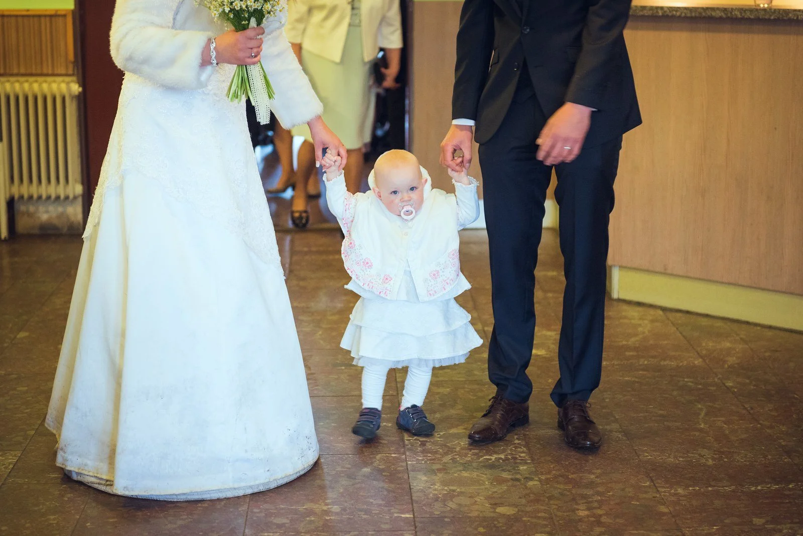 Malá dívka v bílých šatech stojí mezi nevěstou a ženichem u dřevěné stěny.