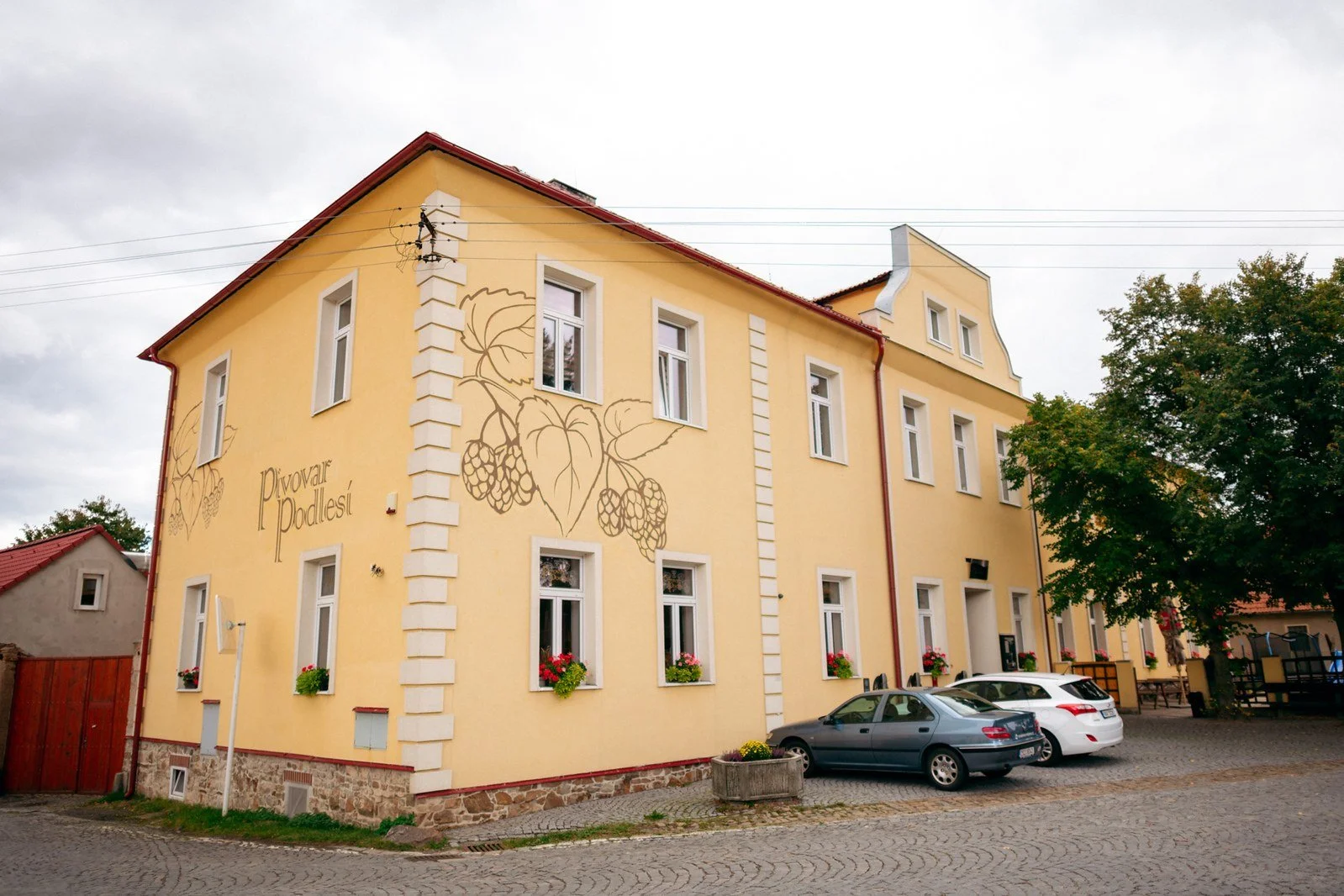 Žlutý rohový dům s malovaným logem a nápisem stojí u parkoviště pod zataženou oblohou.