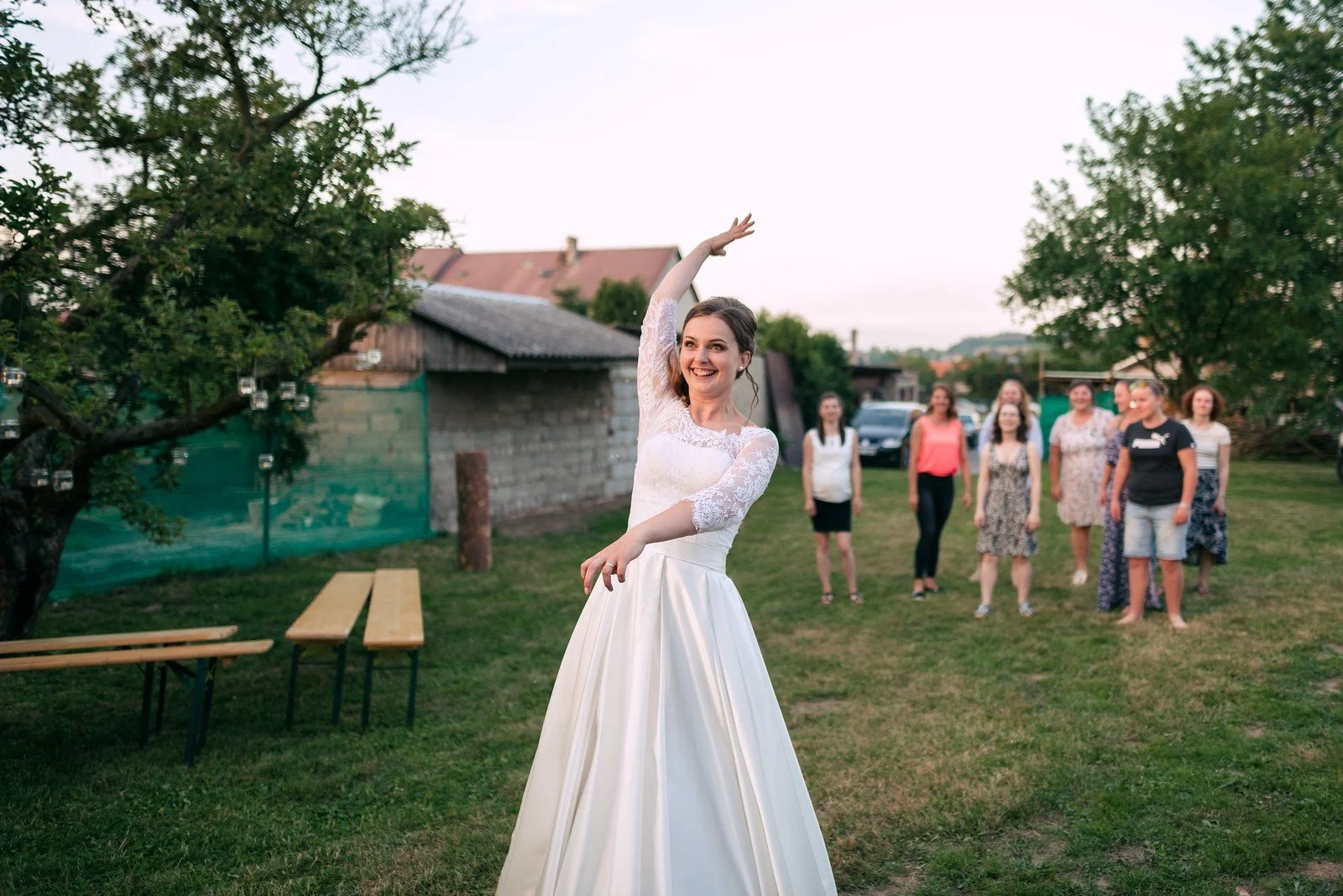 Žena v bílých šatech zvedá ruce zády k hostům na trávníku.