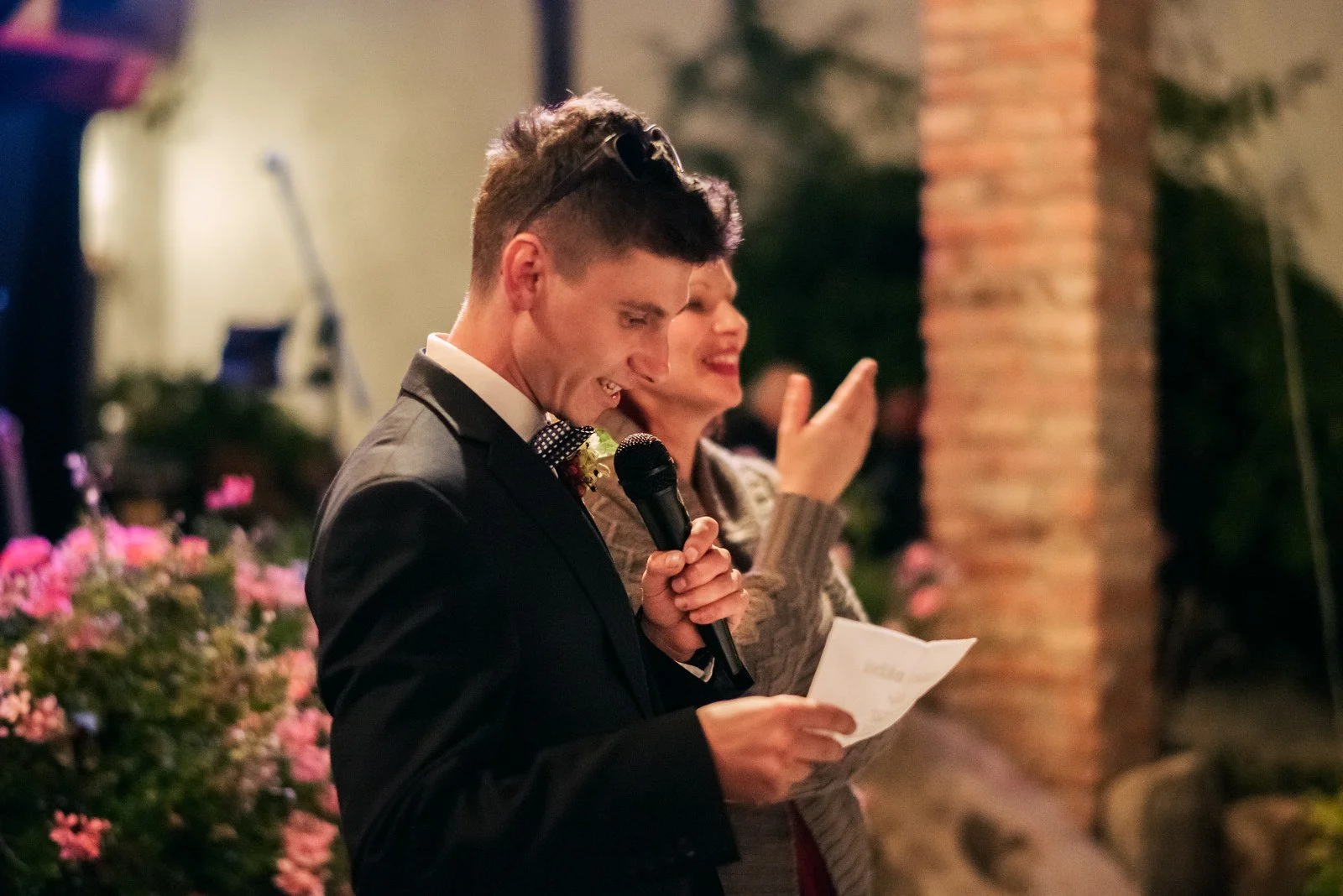 Muž v obleku čte z papíru do mikrofonu u stolu s květinami.