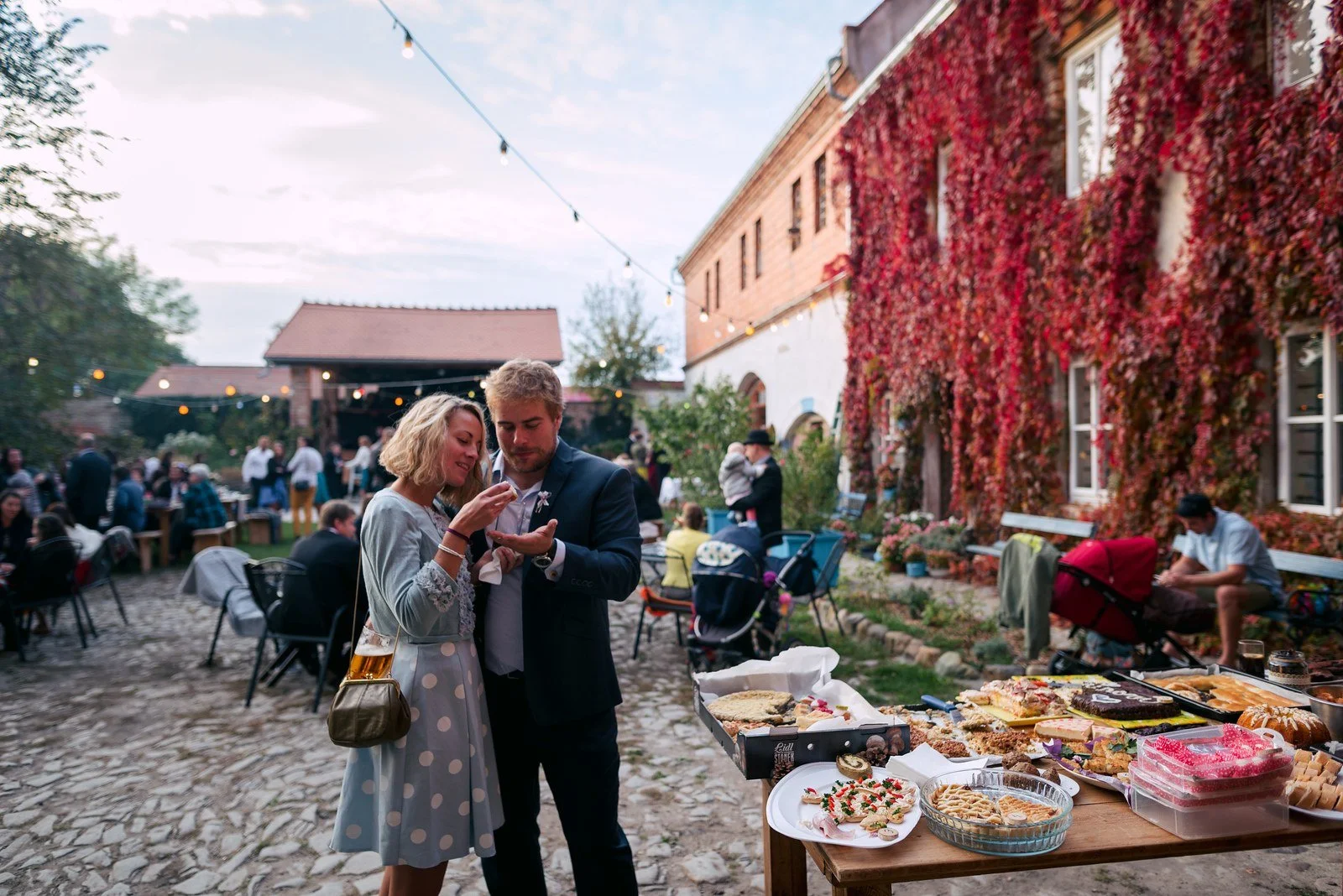 Muž v obleku a žena stojí vedle stolu s občerstvením na večerním nádvoří.