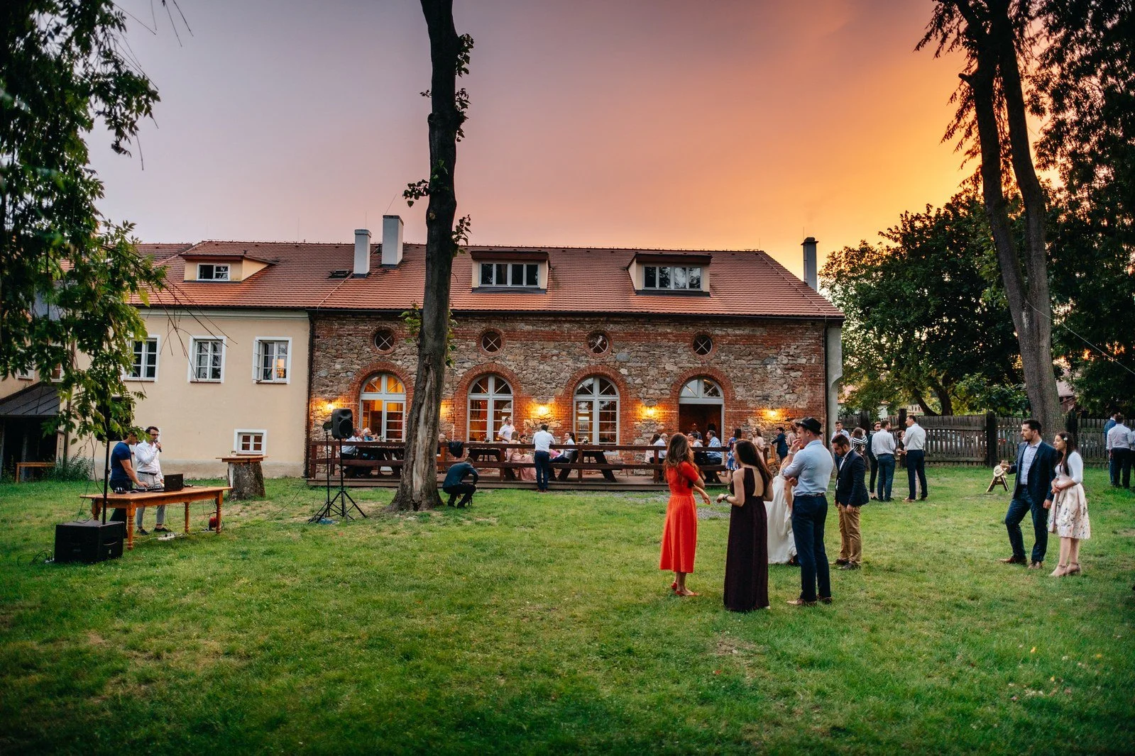 Večerní pohled na zahradu a nasvícenou budovu pod barevnou oblohou.