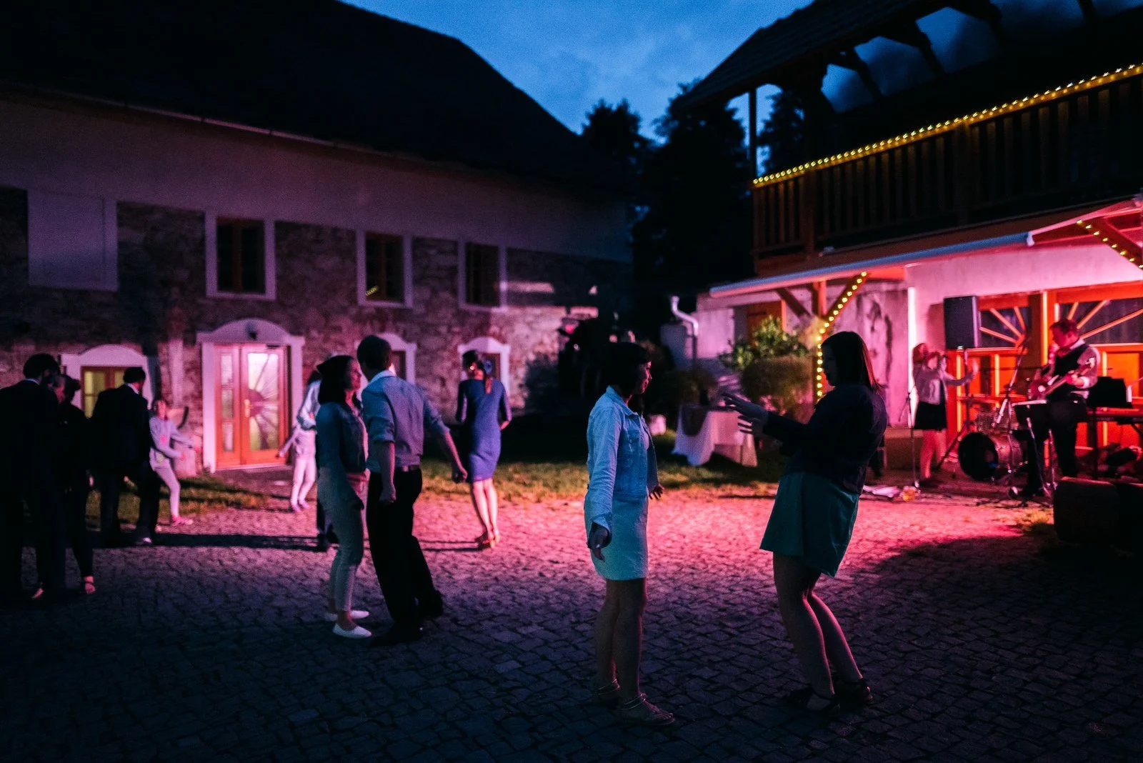 Lidé tančí po setmění na dvoře osvětleném modrými, růžovými a oranžovými světly.