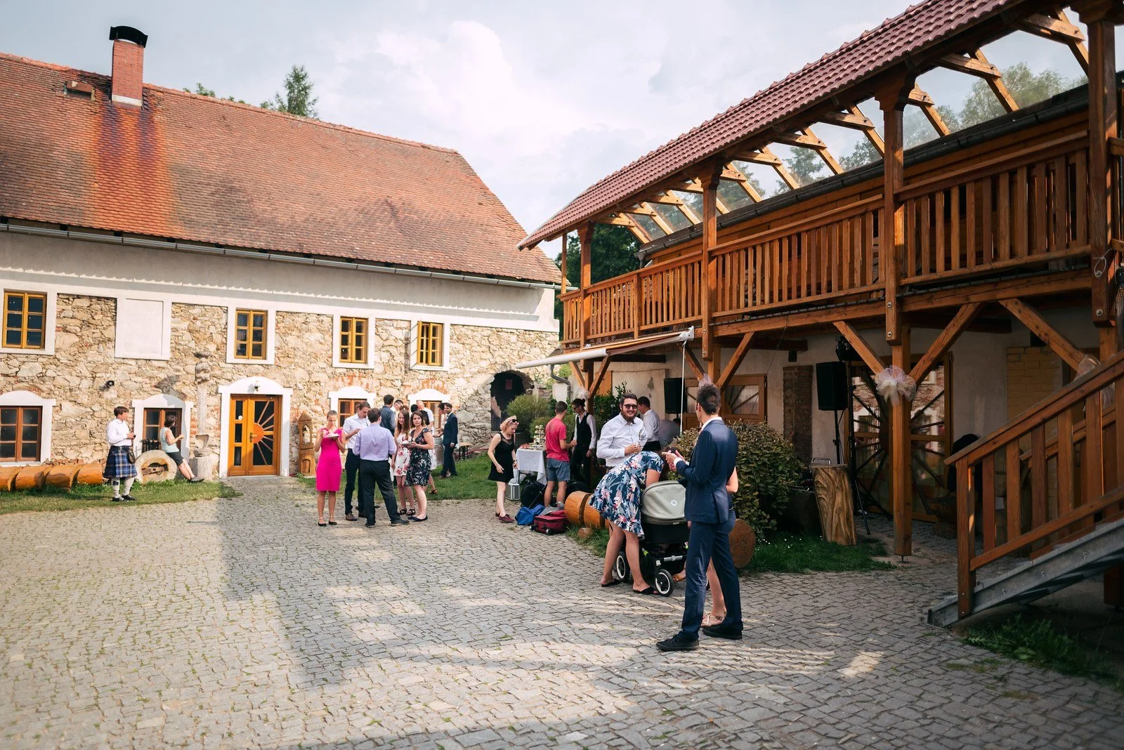 Široký pohled ukazuje kamenné nádvoří s lidmi před budovami a dřevěnou pavlačí.