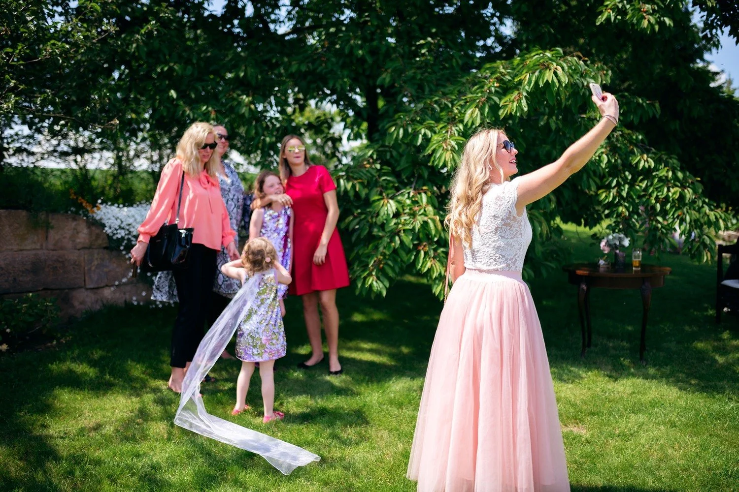 Žena v dlouhé růžové sukni si na zahradě pořizuje selfie, zatímco si dvě malé dívky hrají s bílým závojem.
