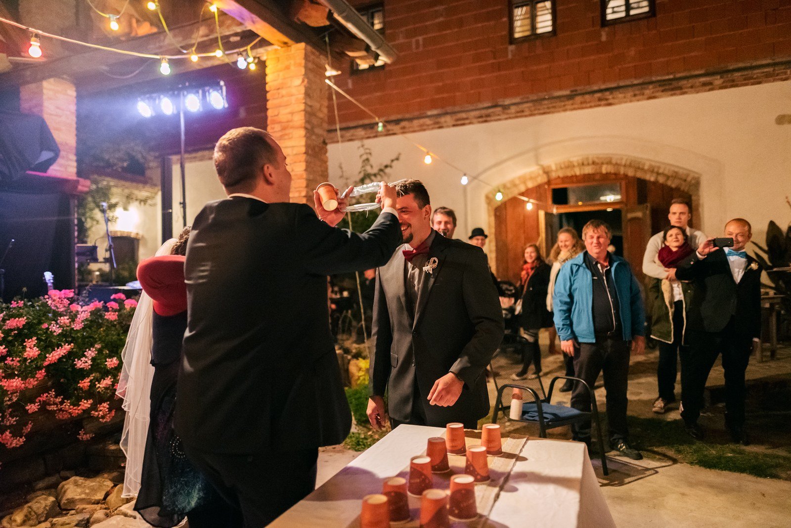Dva muži soutěží u stolu s dřevěnými válečky, zatímco hosté sledují.