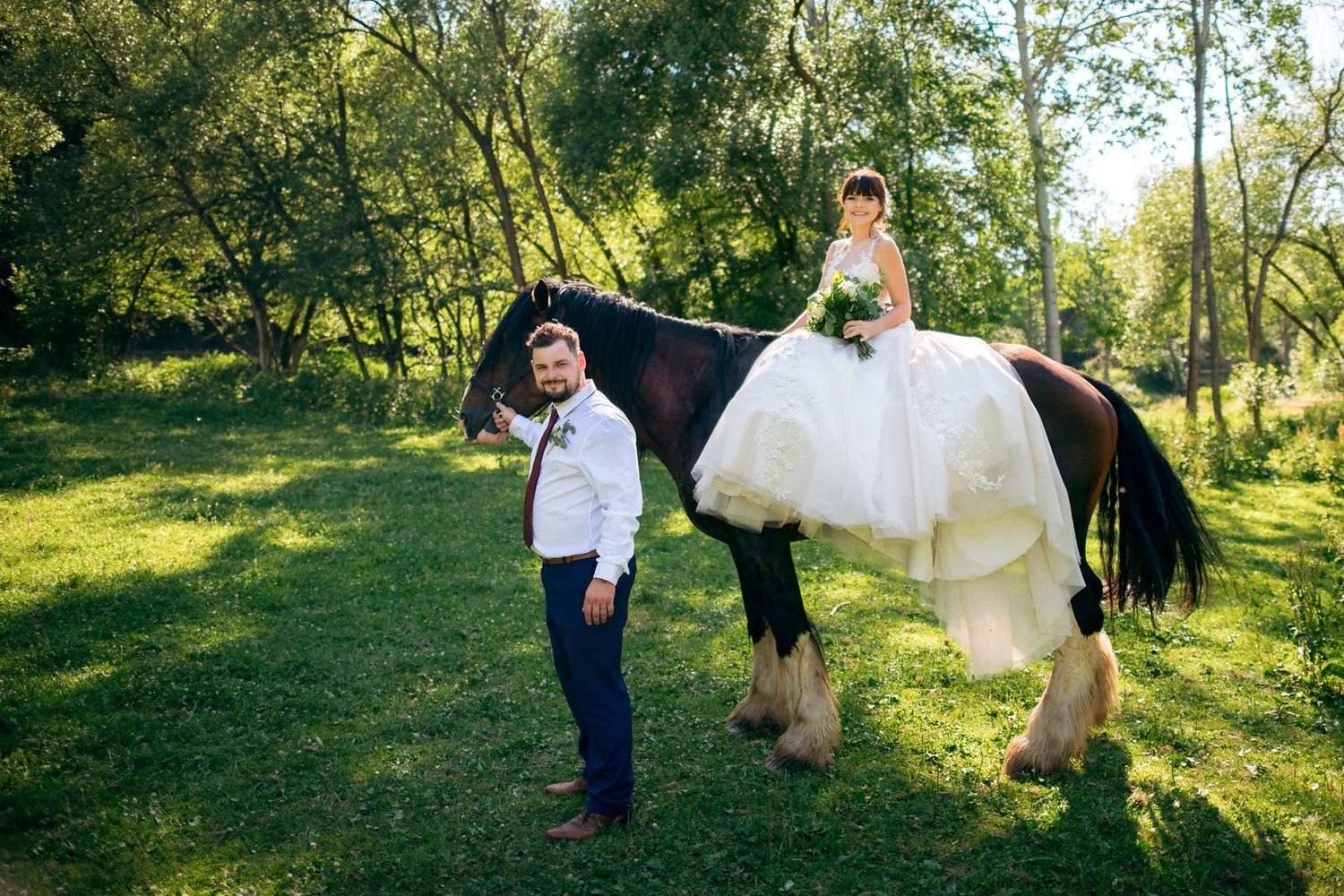 Radka sedí ve svatebních šatech na koni a Pavel stojí vedle něj na louce.