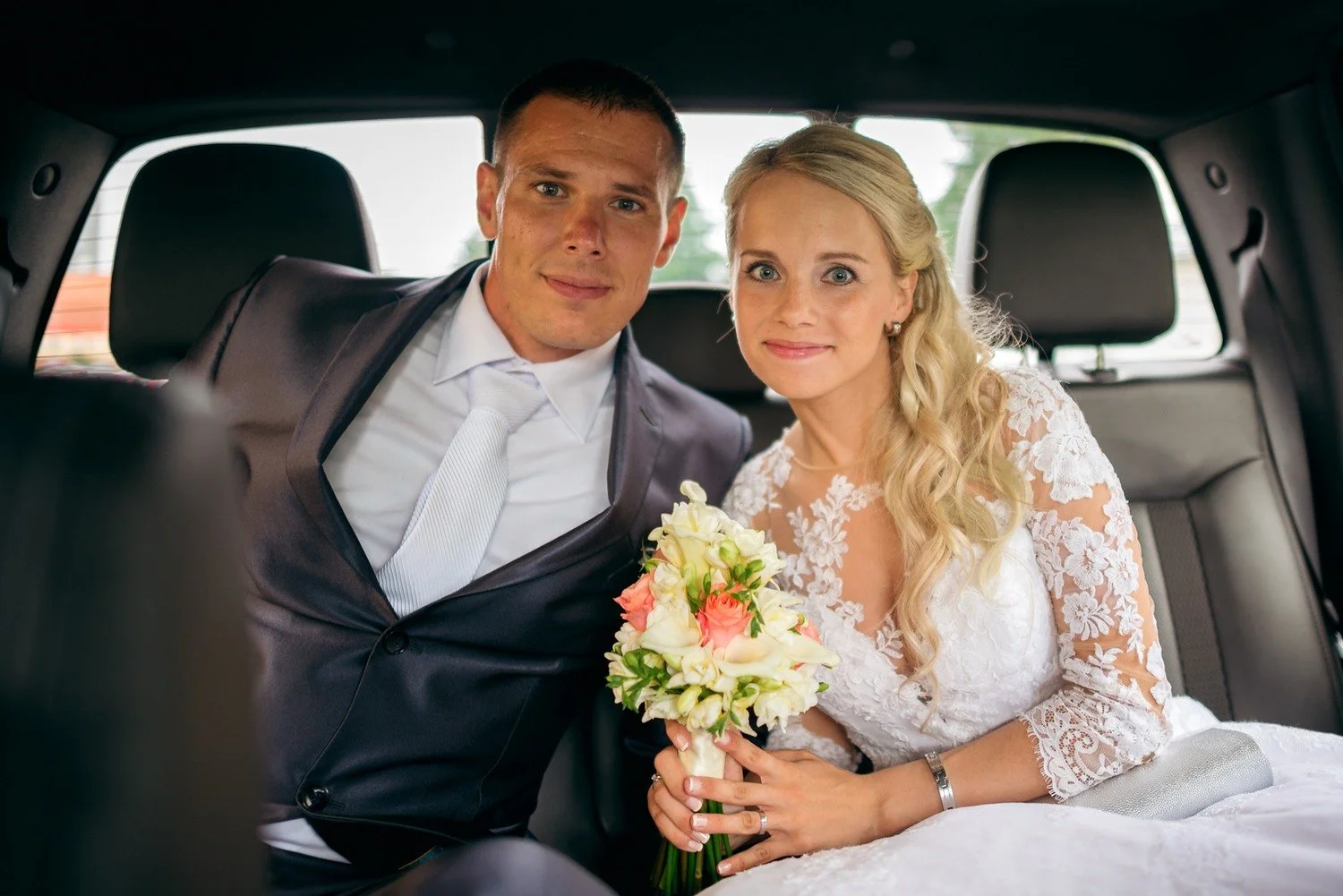 Jitka s kyticí a Lukáš sedí těsně vedle sebe na zadním sedadle auta a hledí do fotoaparátu.