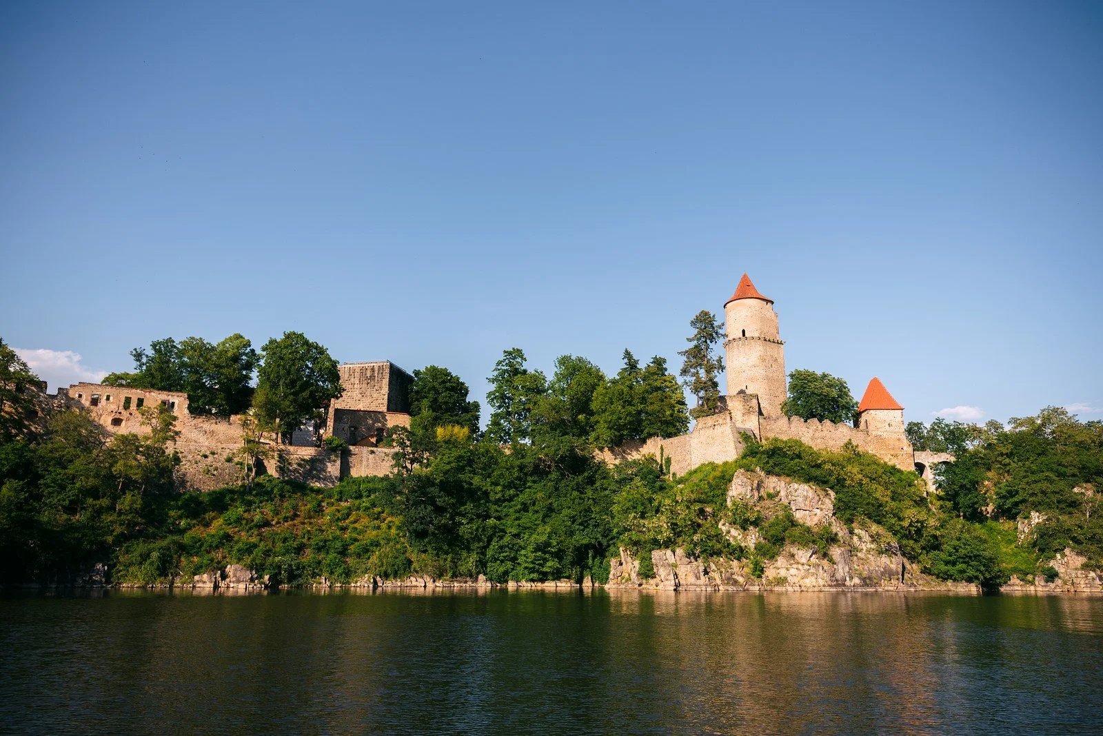 Historická stavba na zalesněném břehu se zrcadlí v modré hladině.