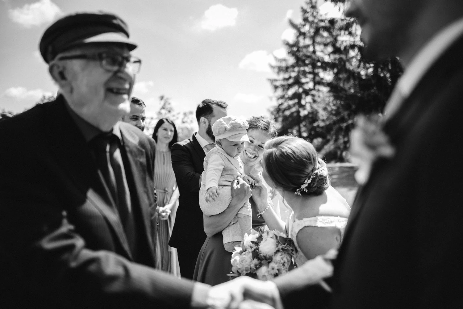 Černobílá momentka nevěsty naklánějící se k miminku v náručí ženy mezi gratulujícími hosty.
