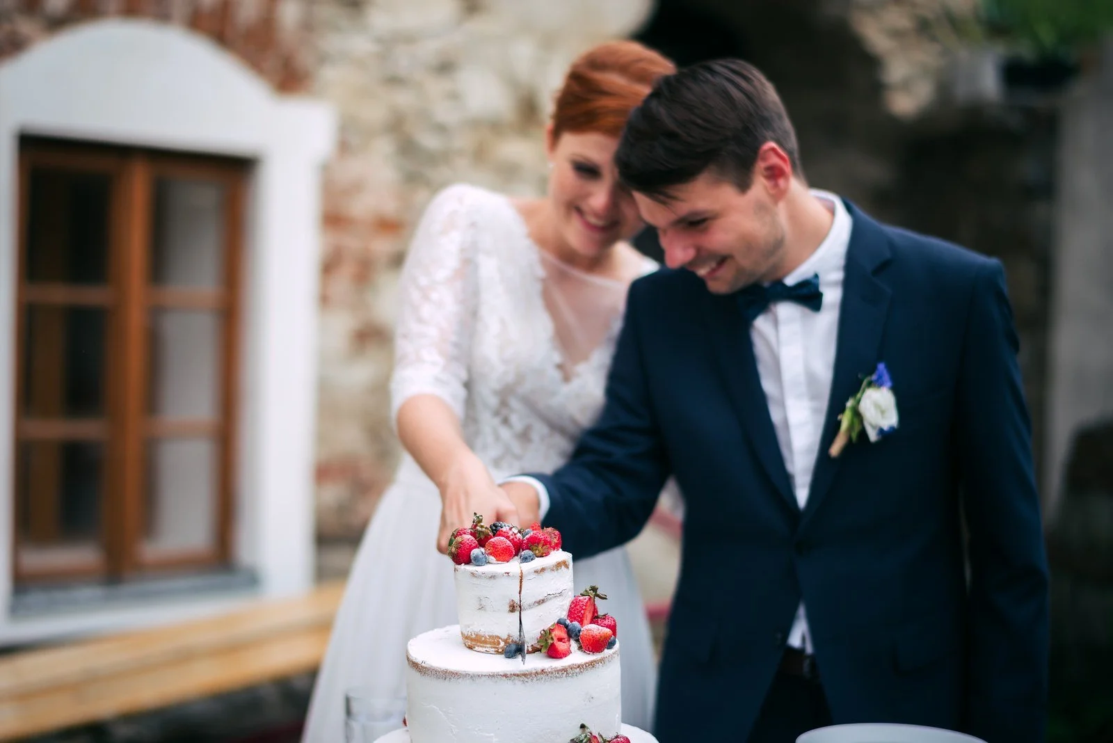 Žena a muž společně krájejí květinami zdobený dort před kamennou budovou.