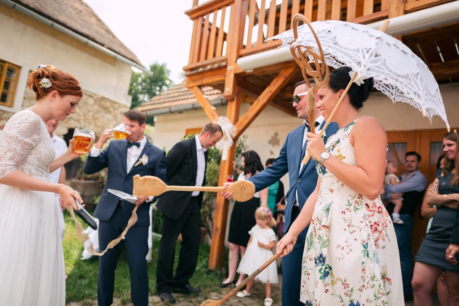 Žena pod bílým krajkovým slunečníkem drží napnutou žlutou stuhu mezi lidmi na nádvoří.