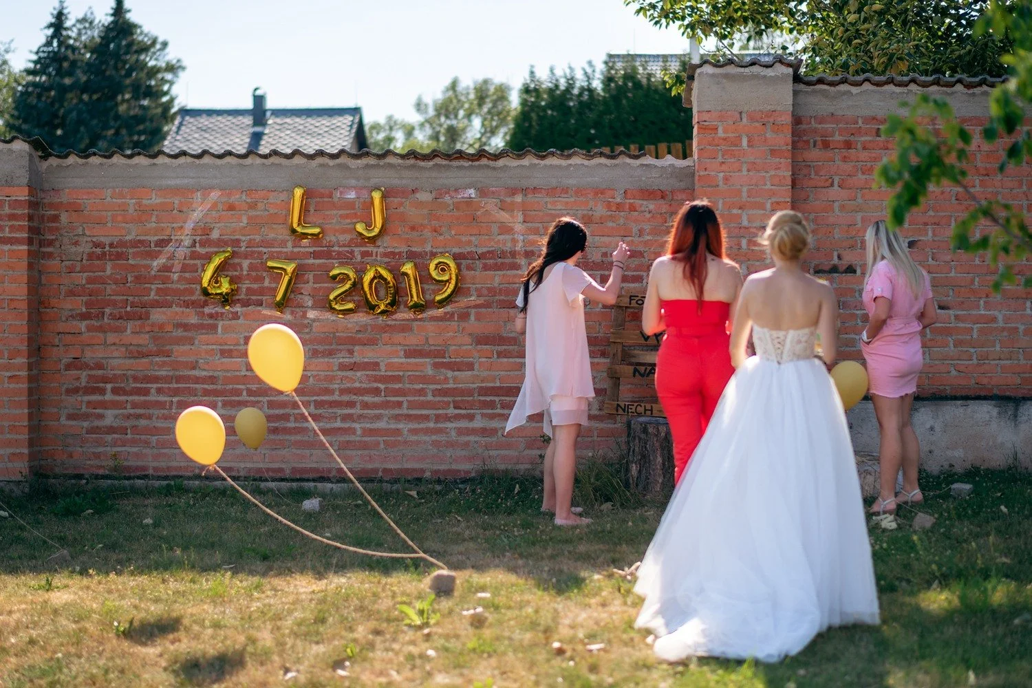Několik žen připevňuje zlaté iniciály a datum na cihlovou zeď, v popředí stojí žena v bílých šatech.