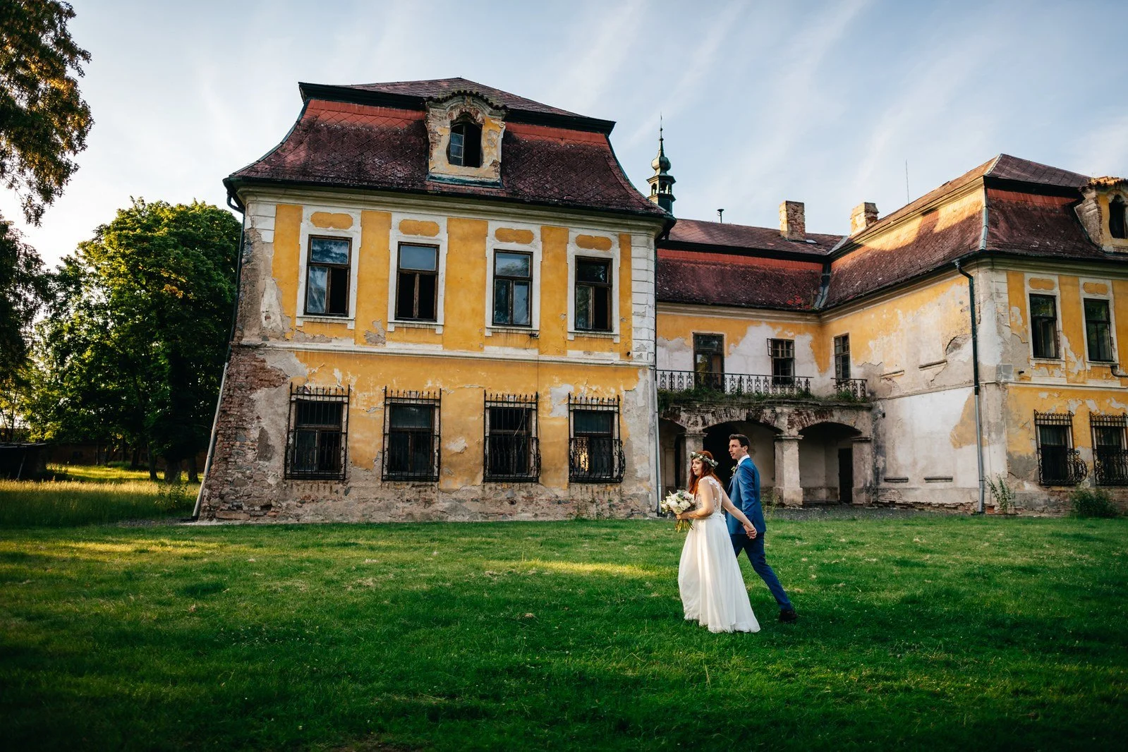 Široký portrét dvojice před historickou budovou a zeleným trávníkem.