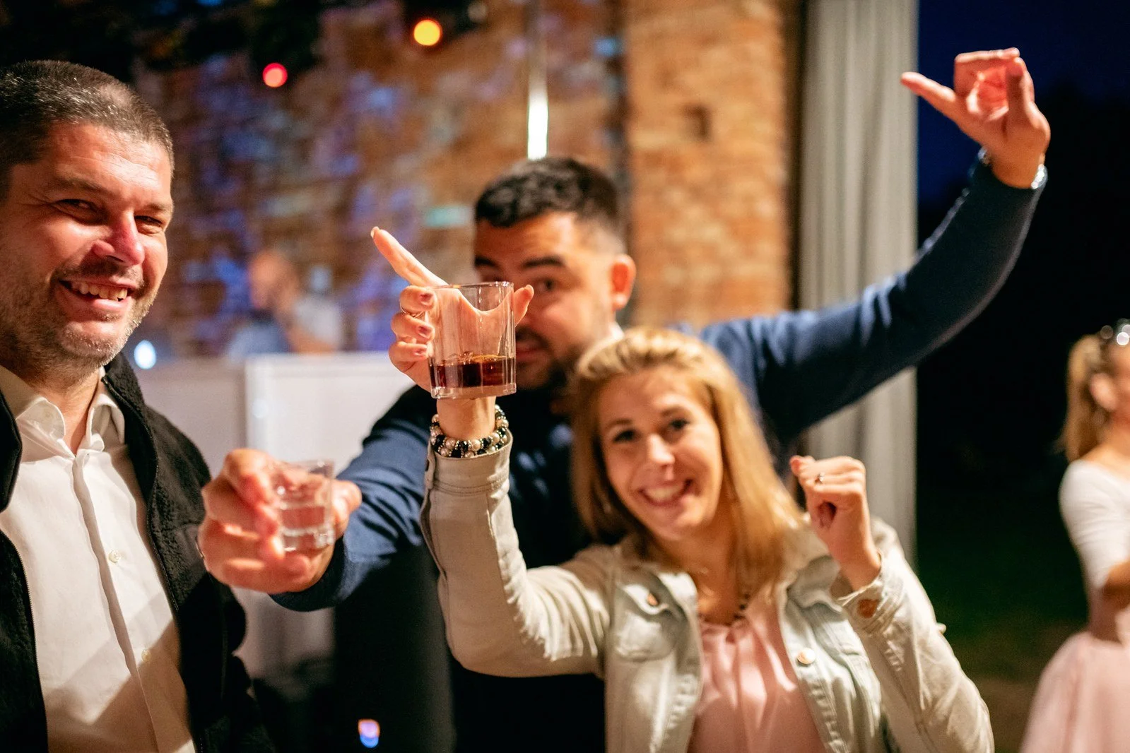 Tři lidé zvedají skleničky směrem k objektivu během večerní zábavy v cihlovém sále.