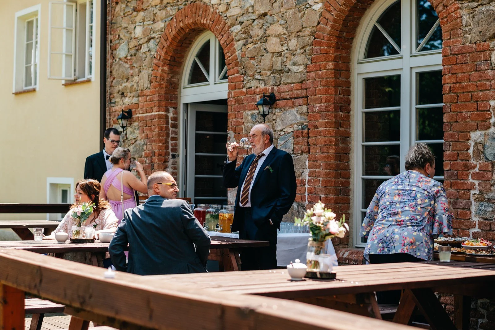 Lidé sedí a stojí u venkovních stolů před cihlovou budovou.