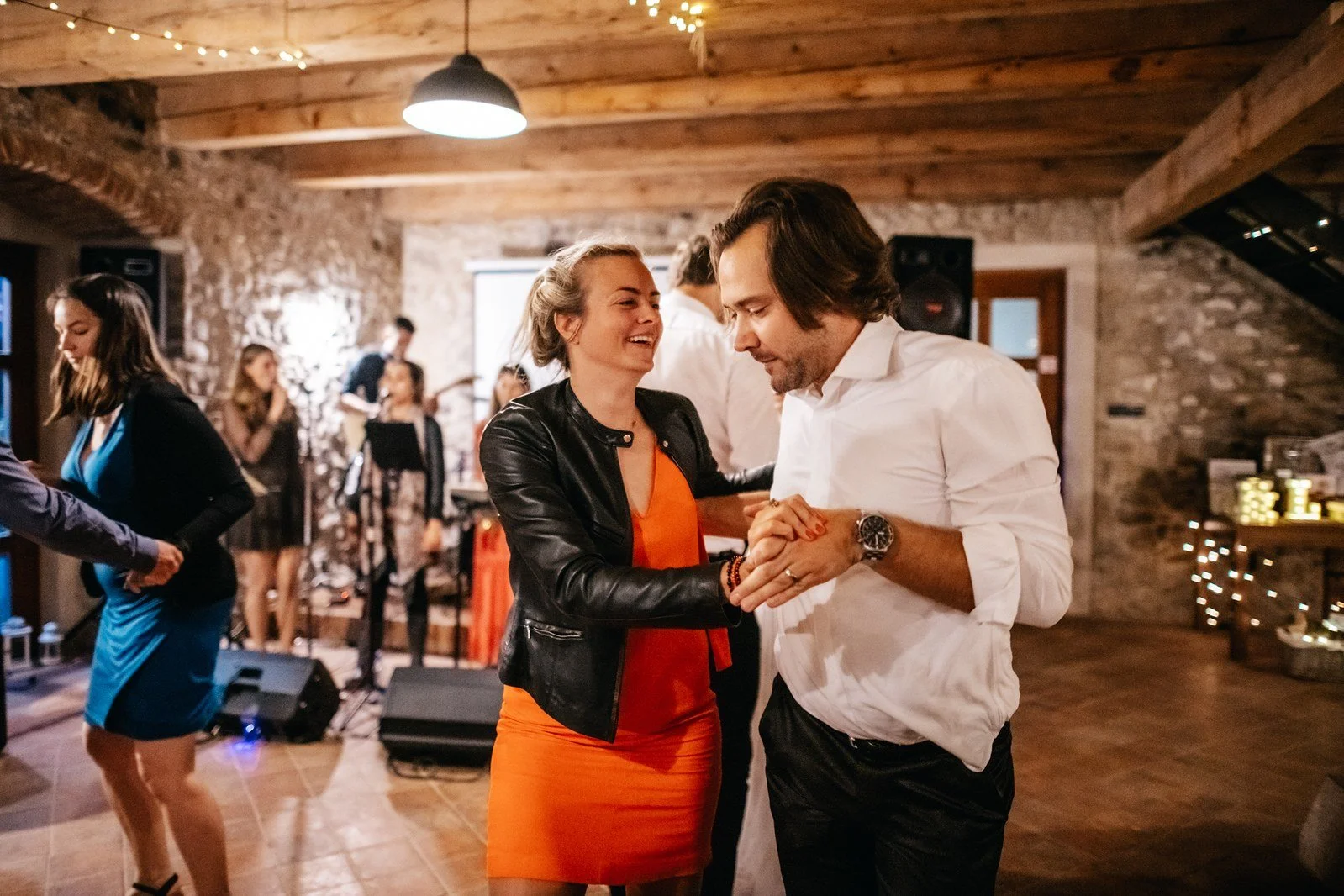 Žena v černém topu a oranžové sukni tančí s mužem v bílé košili pod světelnými řetězy.