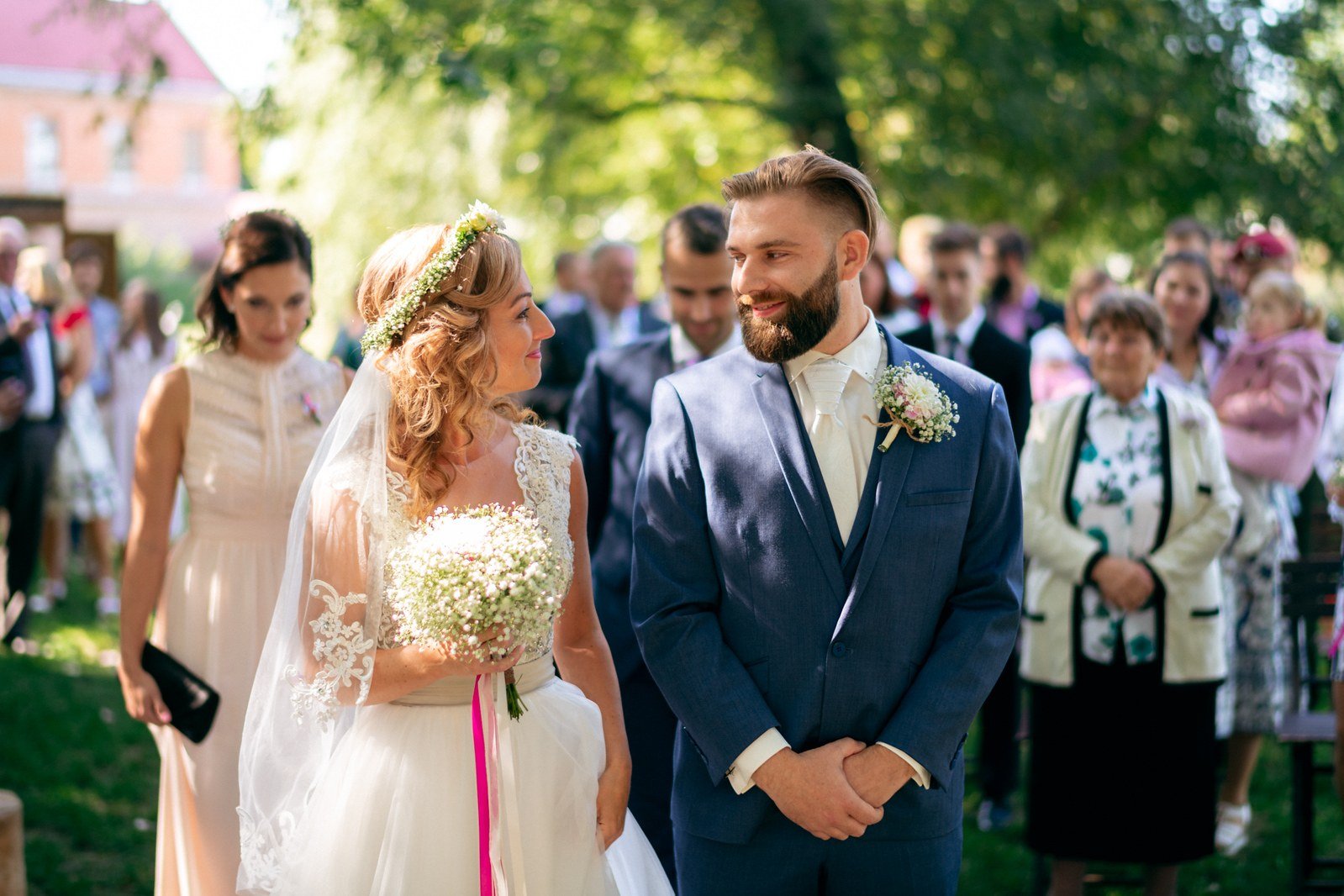 Žena v bílých šatech kráčí zahradní uličkou vedle muže mezi stojícími hosty.