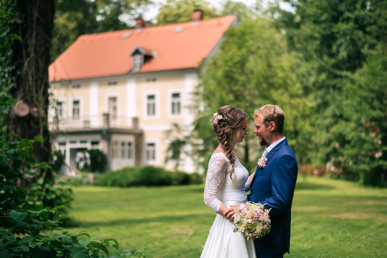 Žena s kyticí a muž v modrém obleku stojí před historickým sídlem.
