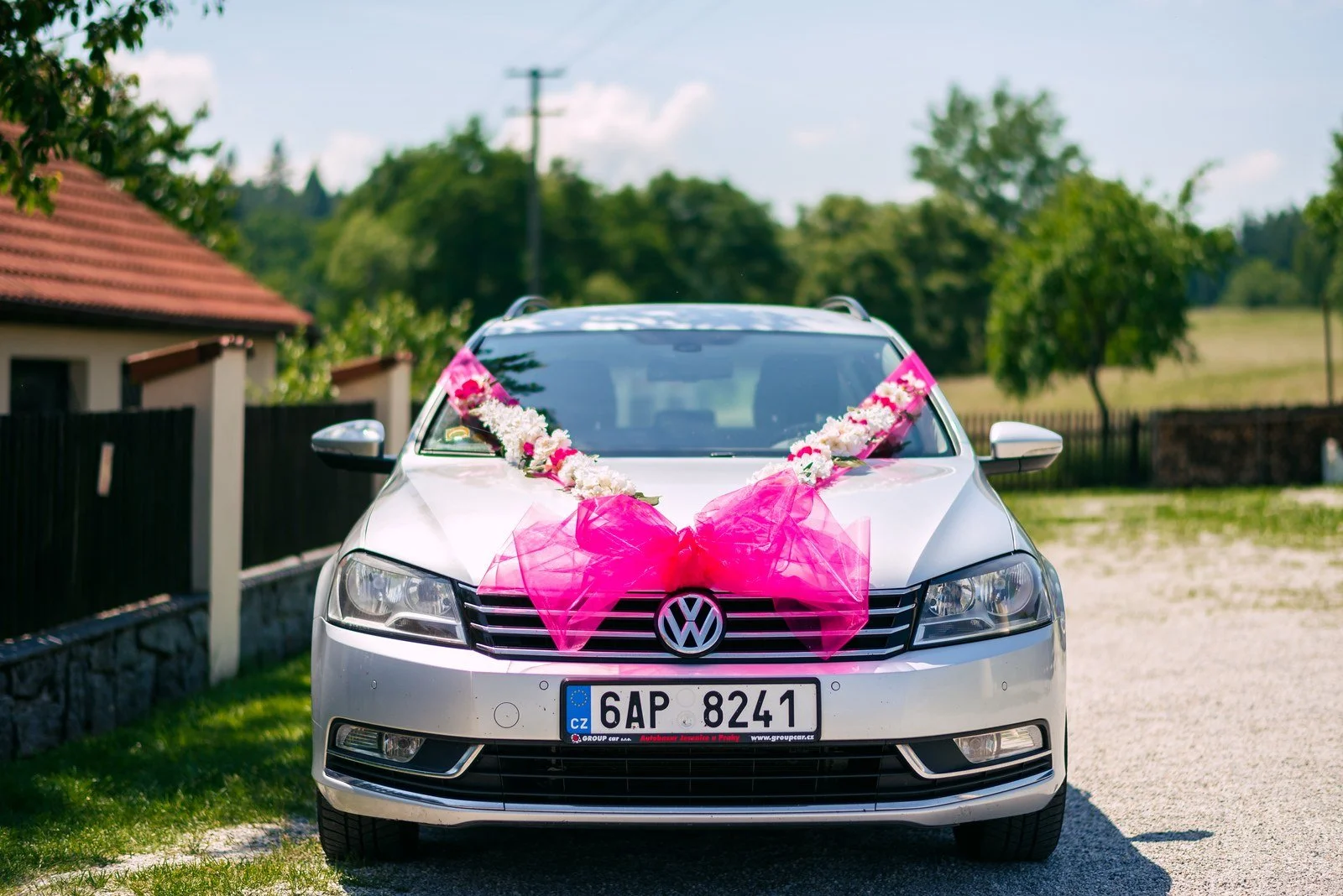 Bílé auto s výraznou růžovou stuhou je připravené před domem.