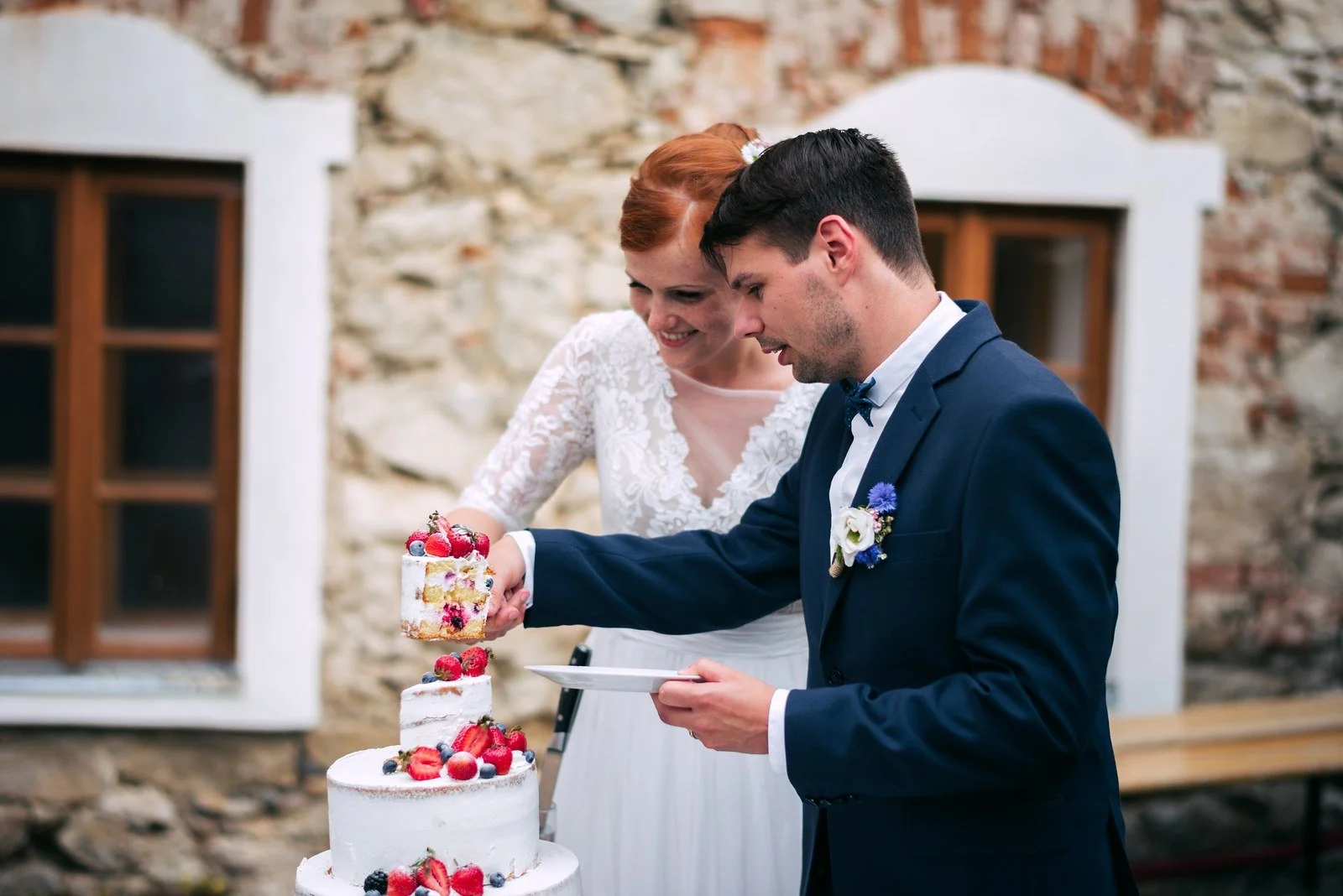 Muž v obleku nabírá porci z bílého patrového dortu, zatímco žena stojí vedle něj.