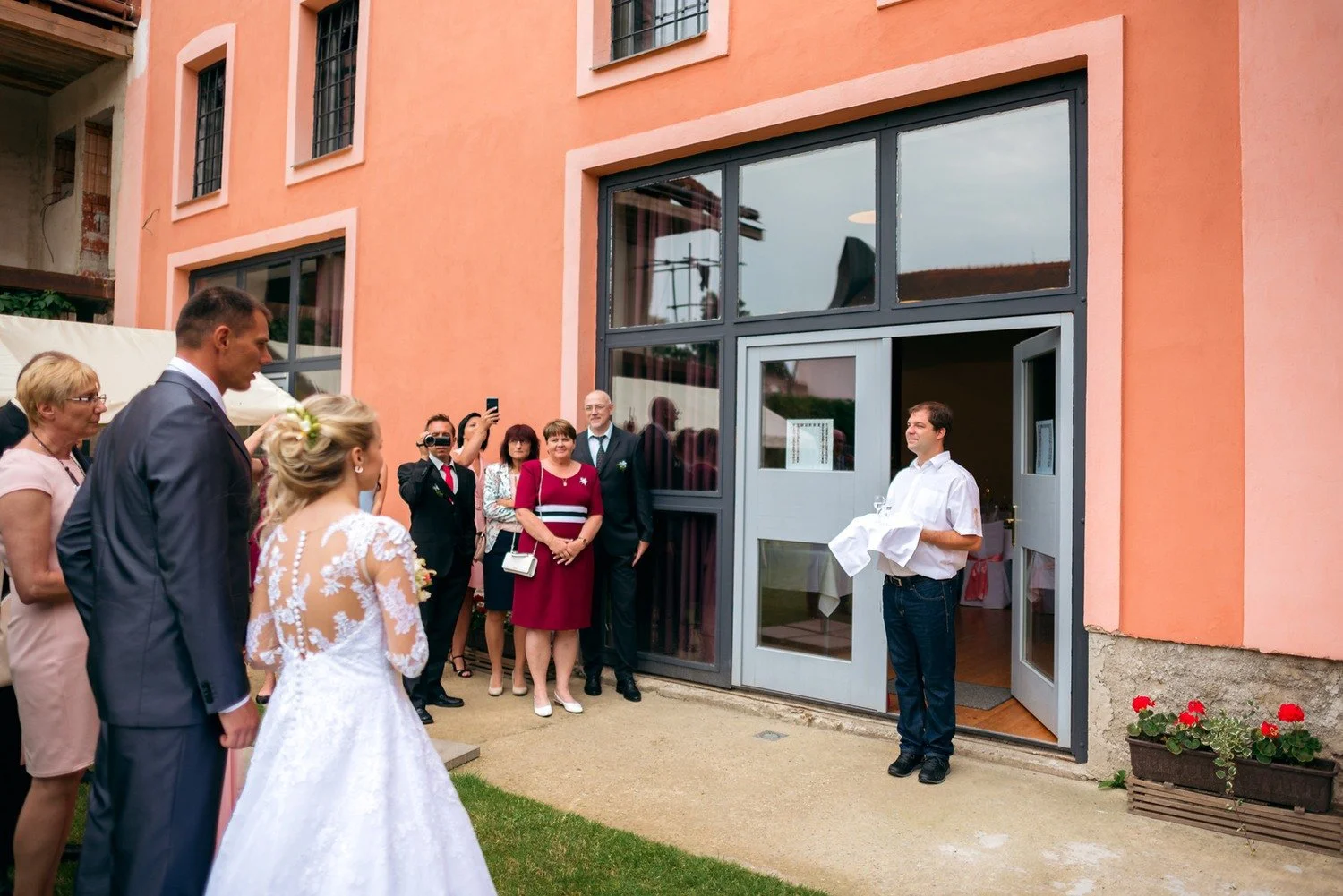 Jitka a Lukáš přicházejí ke vchodu oranžové budovy, před kterým čeká skupina hostů.