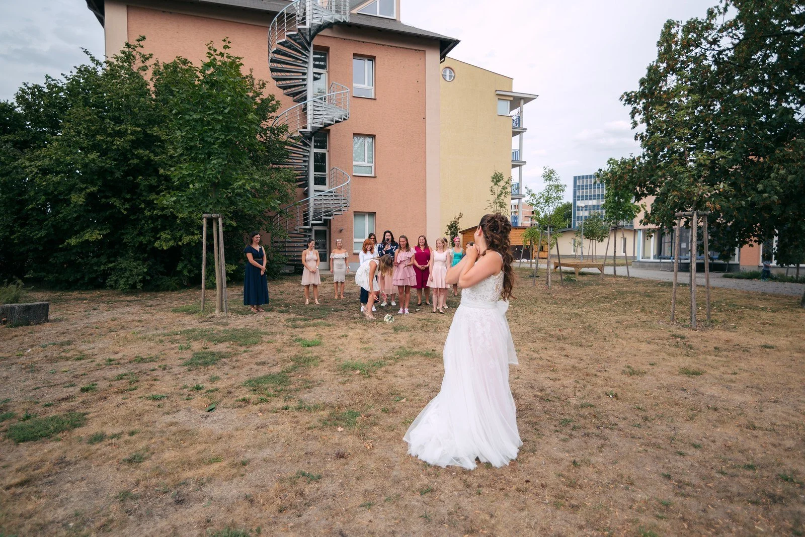 Žena v bílých šatech stojí sama na otevřeném prostranství před růžovou budovou.