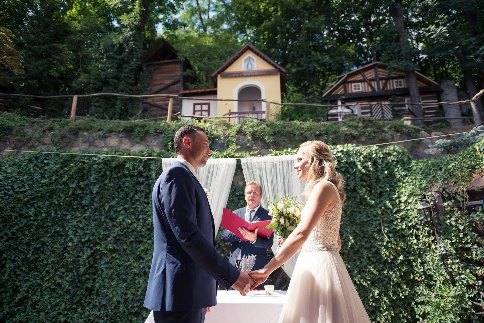 Dvojice si drží ruce před zelenou stěnou a bílou drapérií.