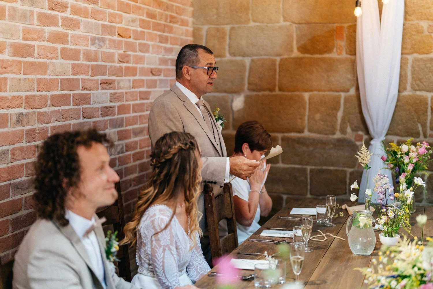 Muž pronáší řeč u svatební tabule, zatímco novomanželé poslouchají