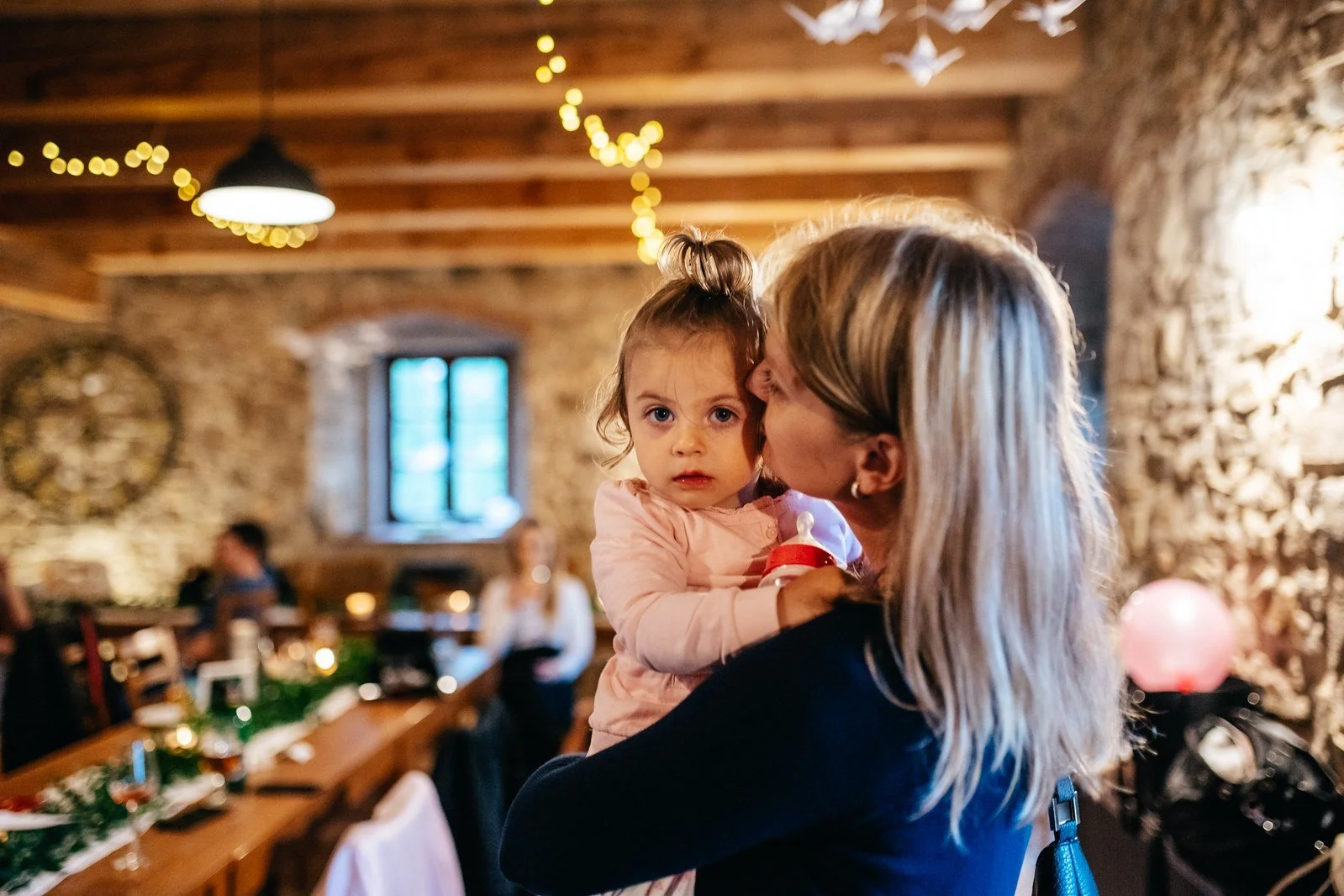 Žena s dlouhými světlými vlasy drží v náručí malé dítě v růžovém oblečení uvnitř sálu.