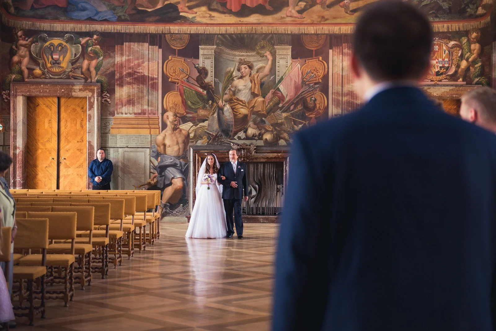 Pohled přes záda muže zachycuje příchod dvojice hluboko v ornamentálním sále.