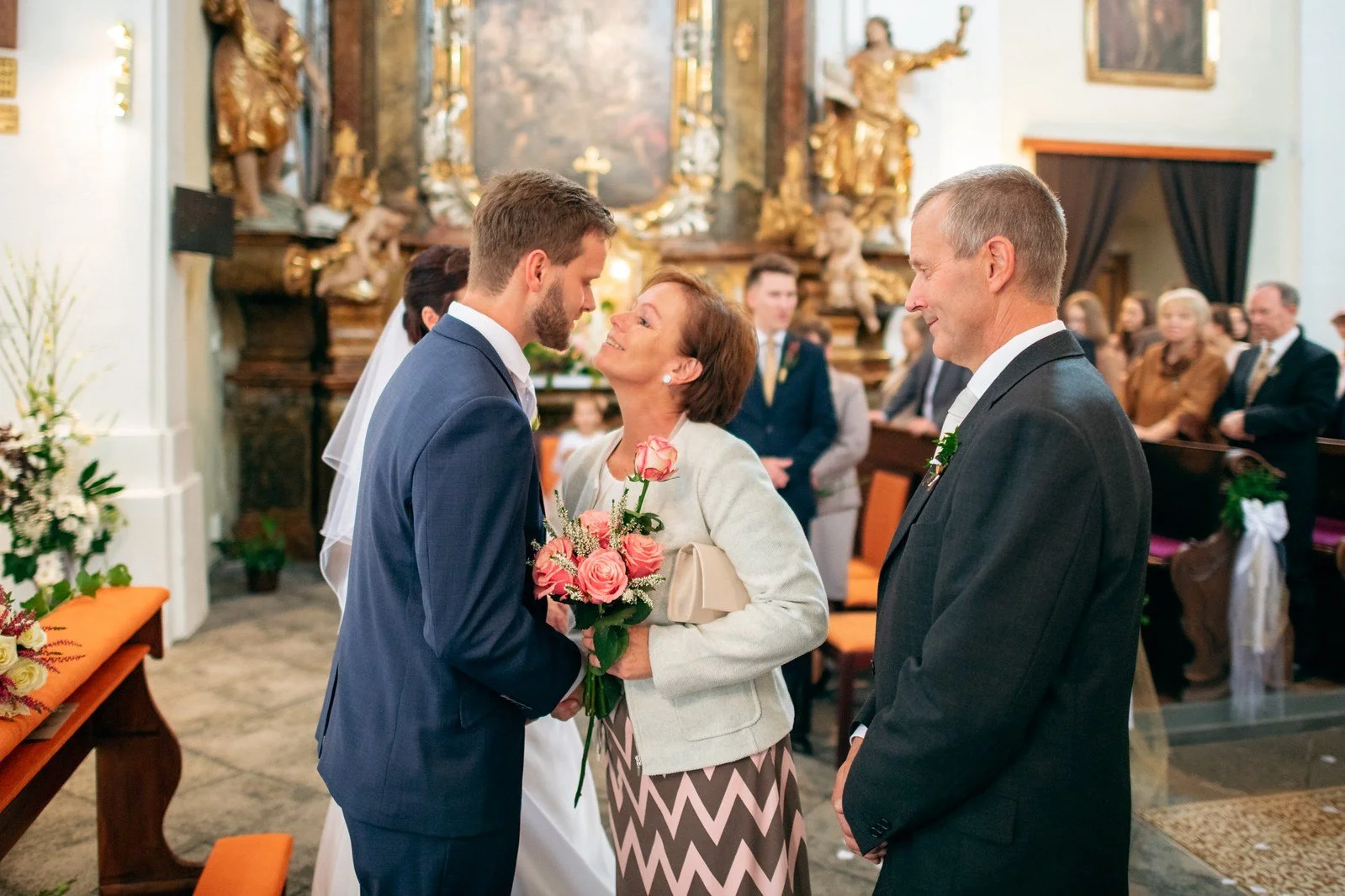 Muž v modrém obleku se naklání k ženě se světlým sakem a růžovou kyticí mezi kostelními hosty.
