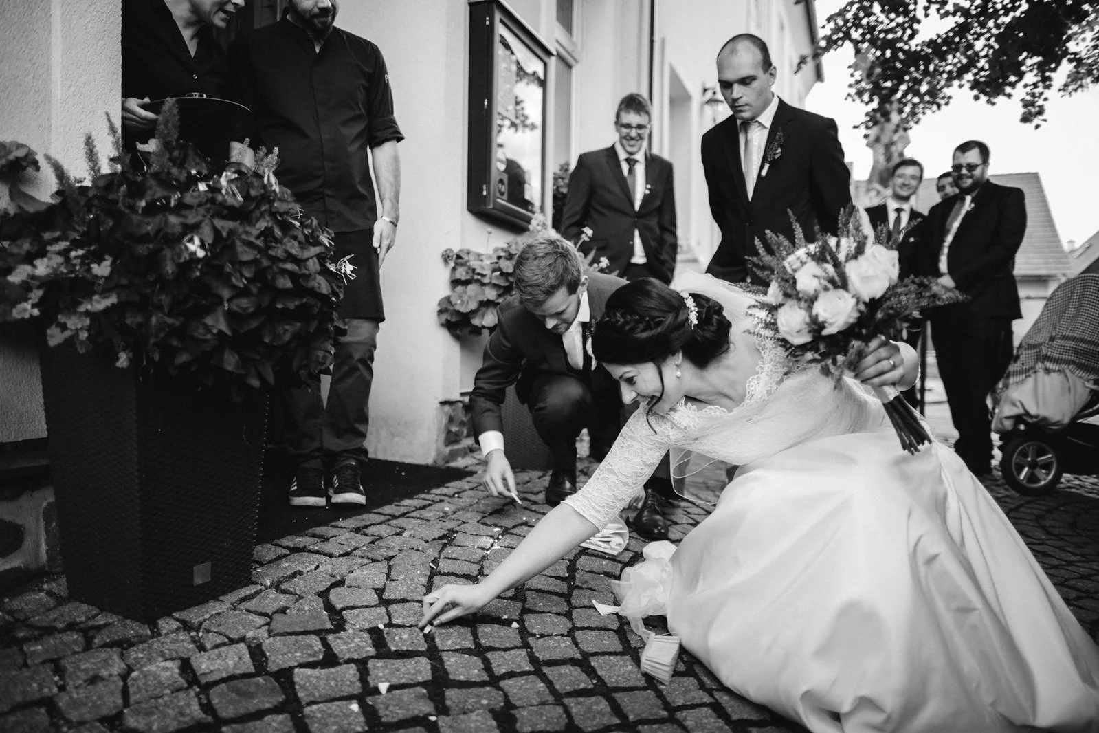 Žena v objemných bílých šatech se v černobílém záběru sklání k dlažbě obklopená několika muži.