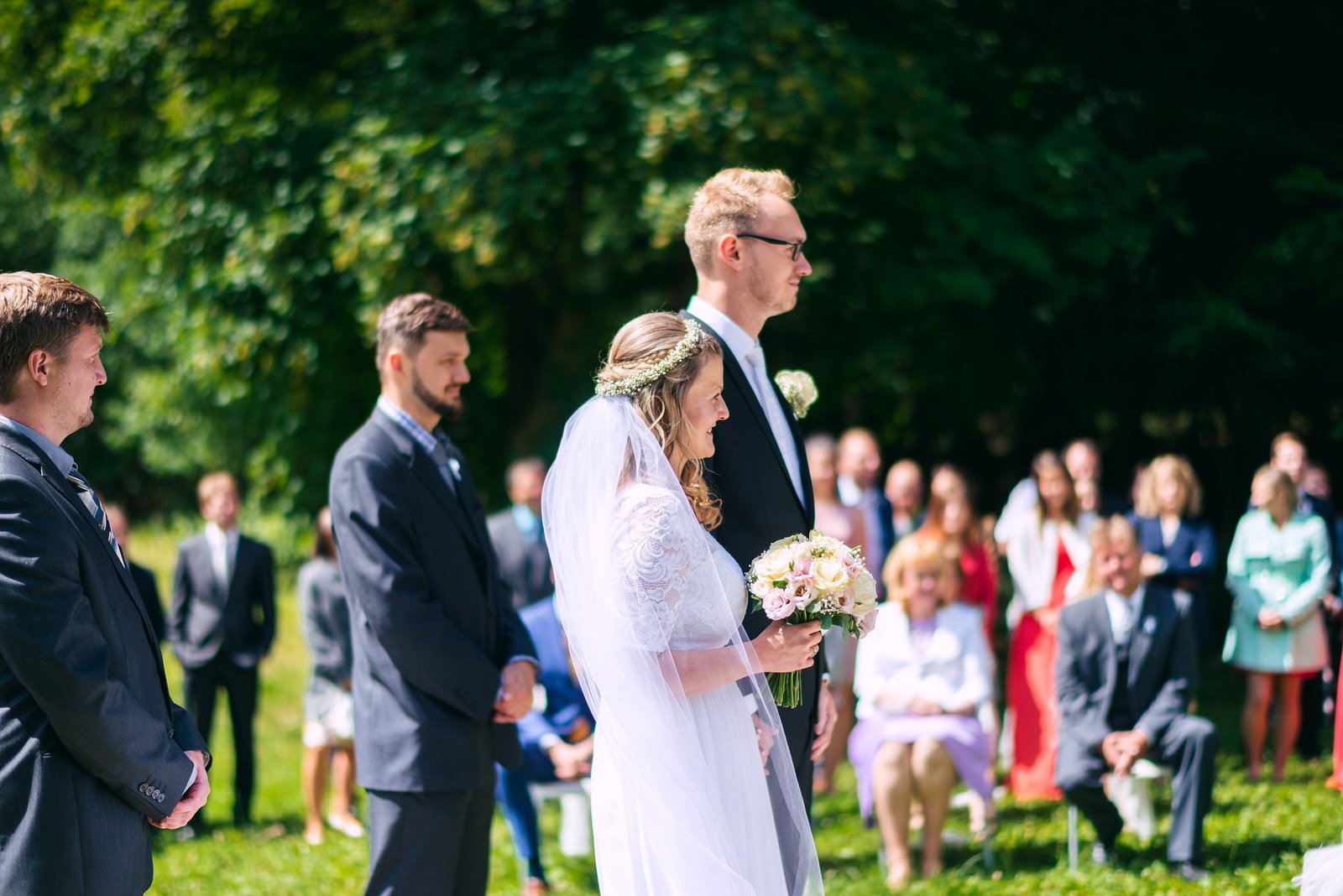 Dva muži v košilích a vestách stojí v pokoji, zatímco žena jednomu upravuje klopu.