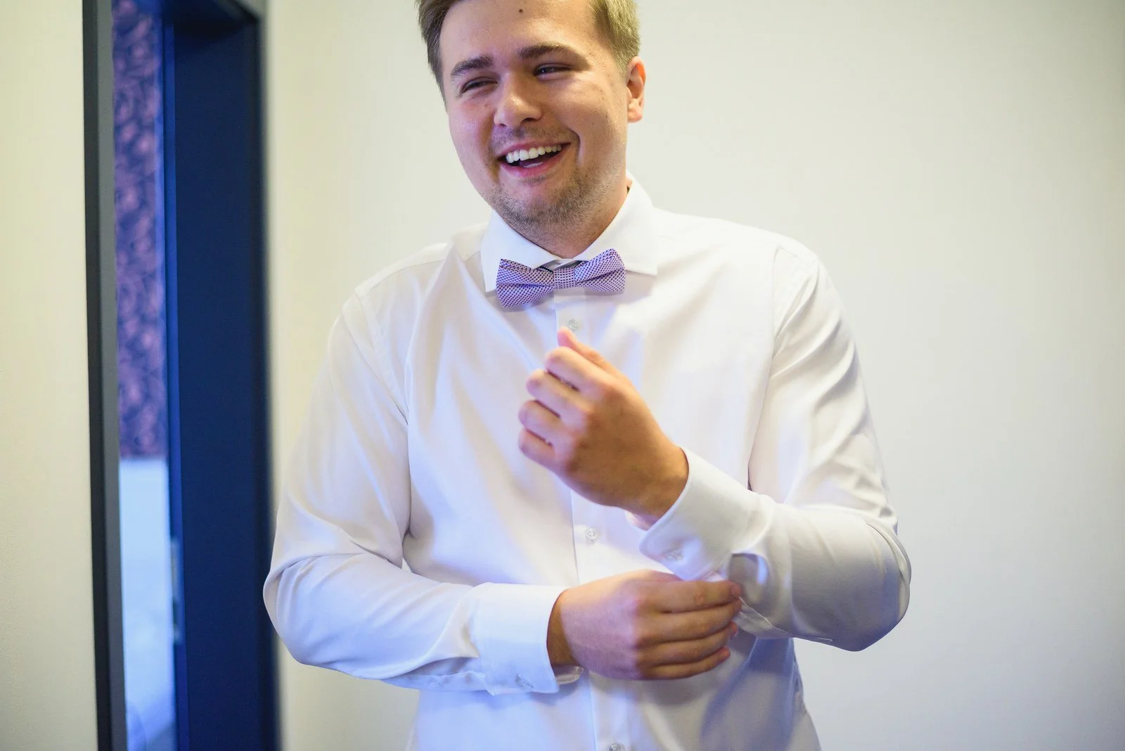 Usměvavý muž v bílé košili si upravuje manžetu rukávu.