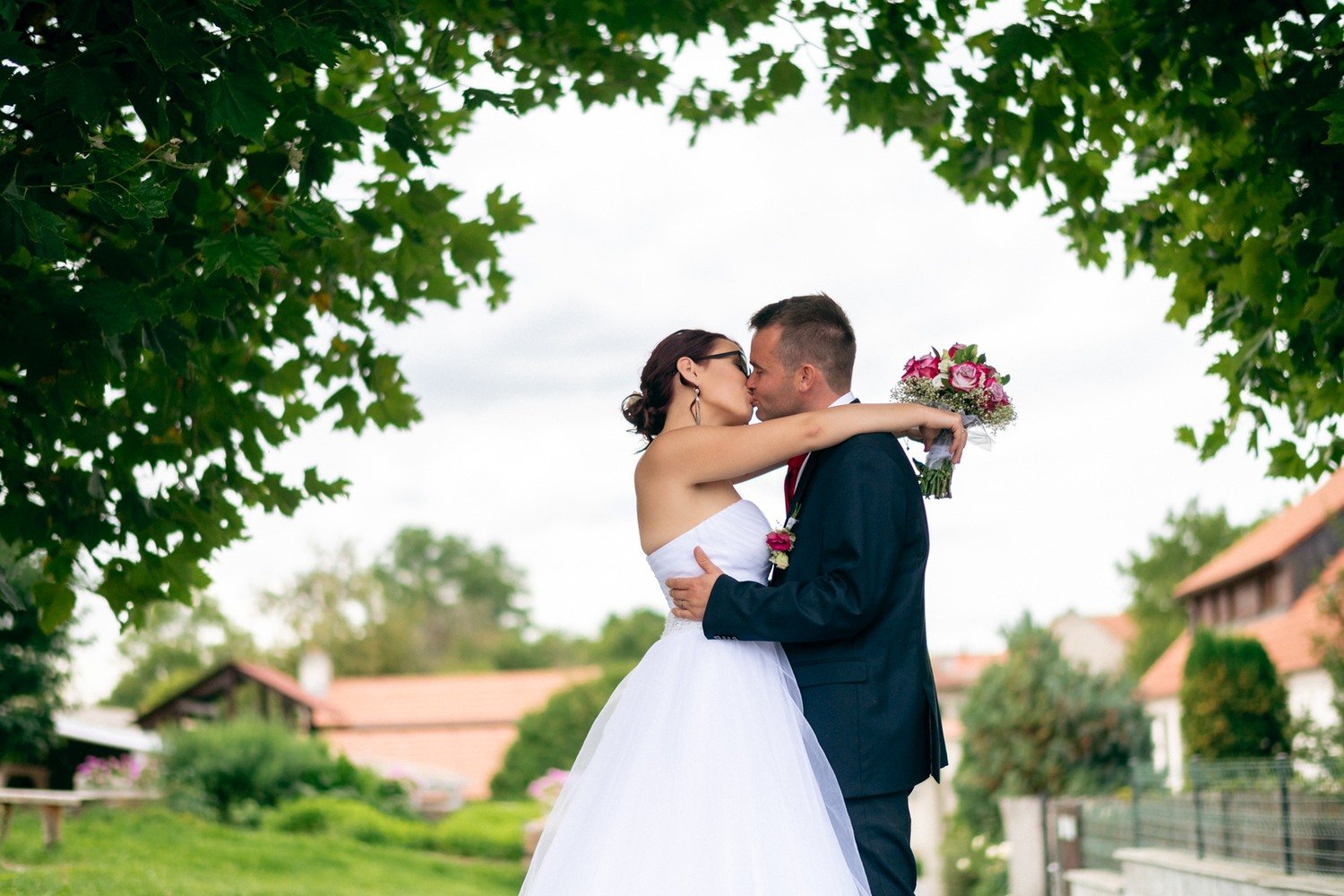 Žena v bílých šatech objímá a líbá muže v obleku pod zelenými větvemi.