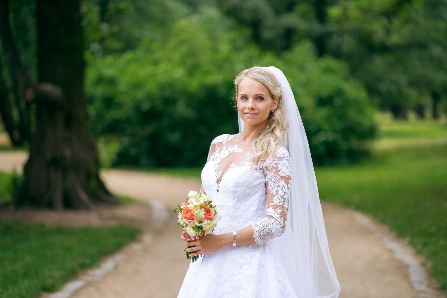 Jitka s kyticí stojí na světlé cestě v parku před rozostřenou zelení.