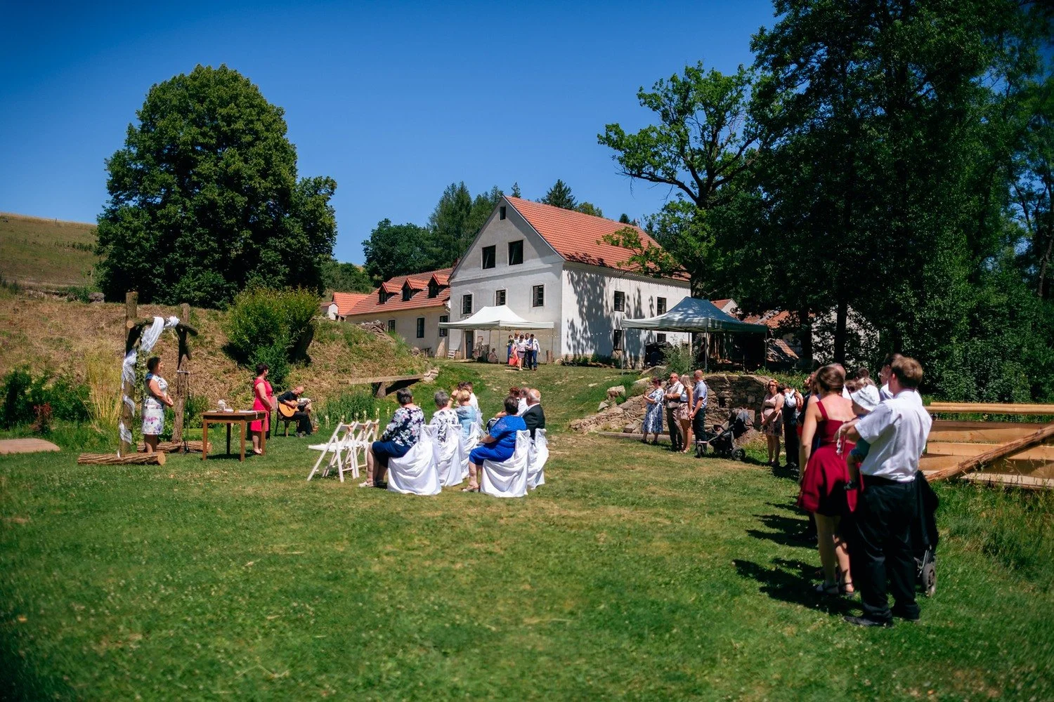 Hosté sedí na bílých židlích před slavobránou, zatímco další přicházejí po trávě.