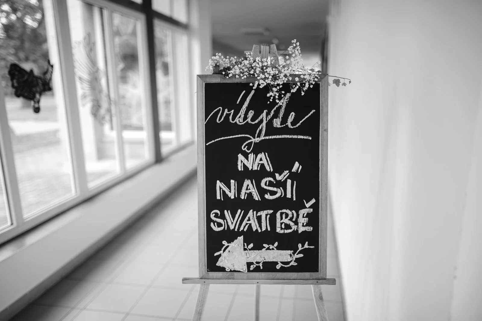 Na okenním parapetu je postavená černá tabule s ručně psaným svatebním nápisem.