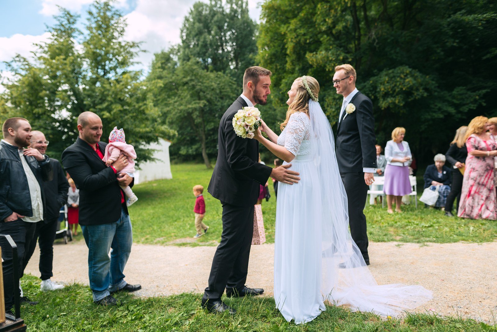 Žena v bílých šatech hovoří venku s mužem v obleku, obklopená dalšími hosty.
