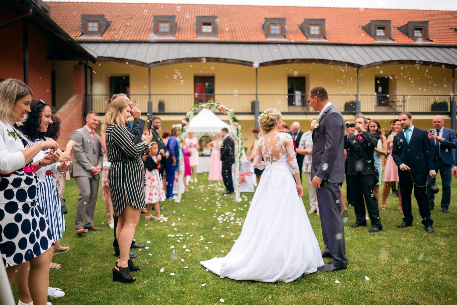 Jitka a Lukáš stojí zády k fotoaparátu uprostřed špalíru hostů a konfet na trávě.