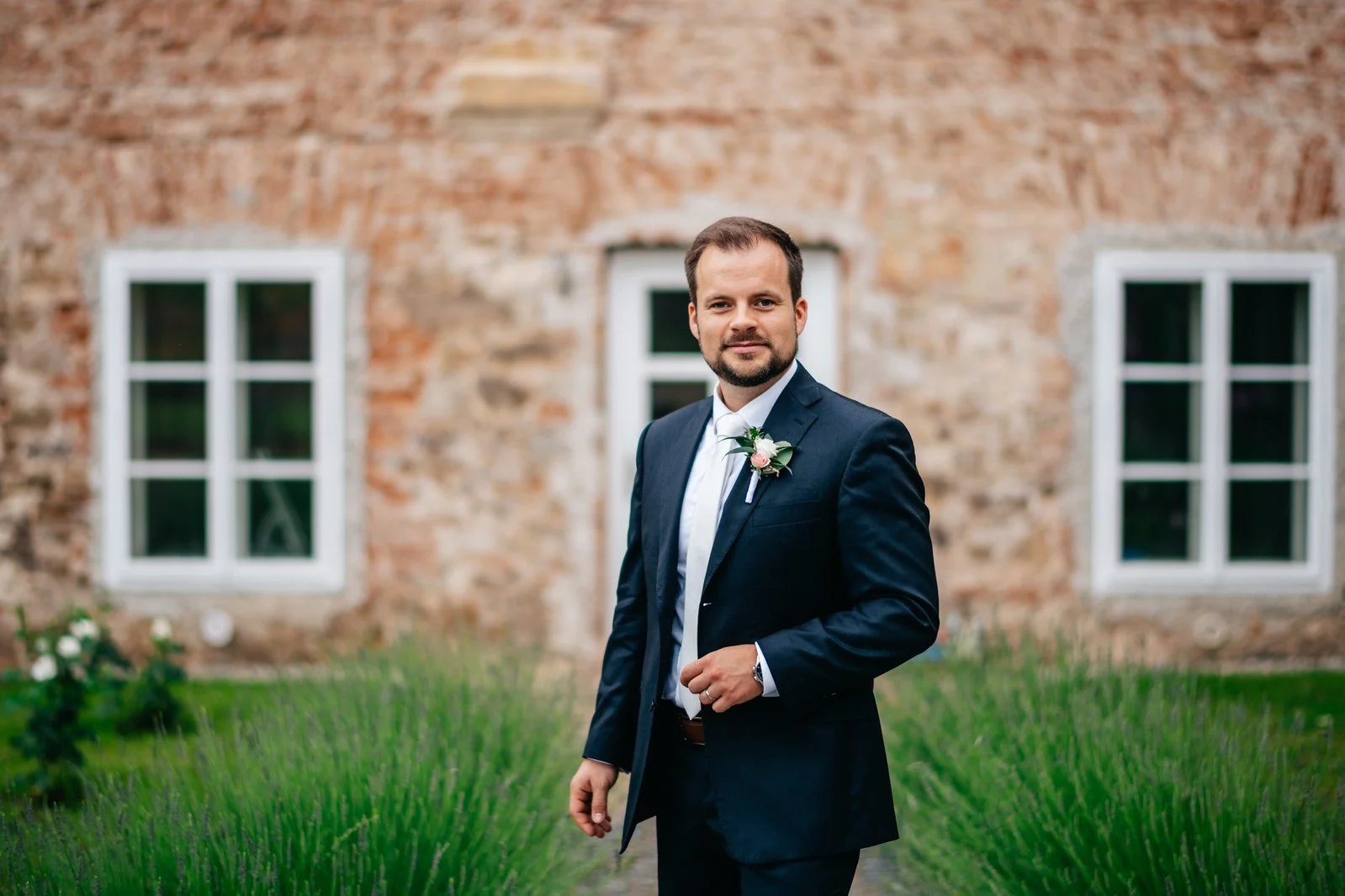 Muž v tmavém obleku stojí s rukou u saka před kamennou fasádou a vysokou trávou.