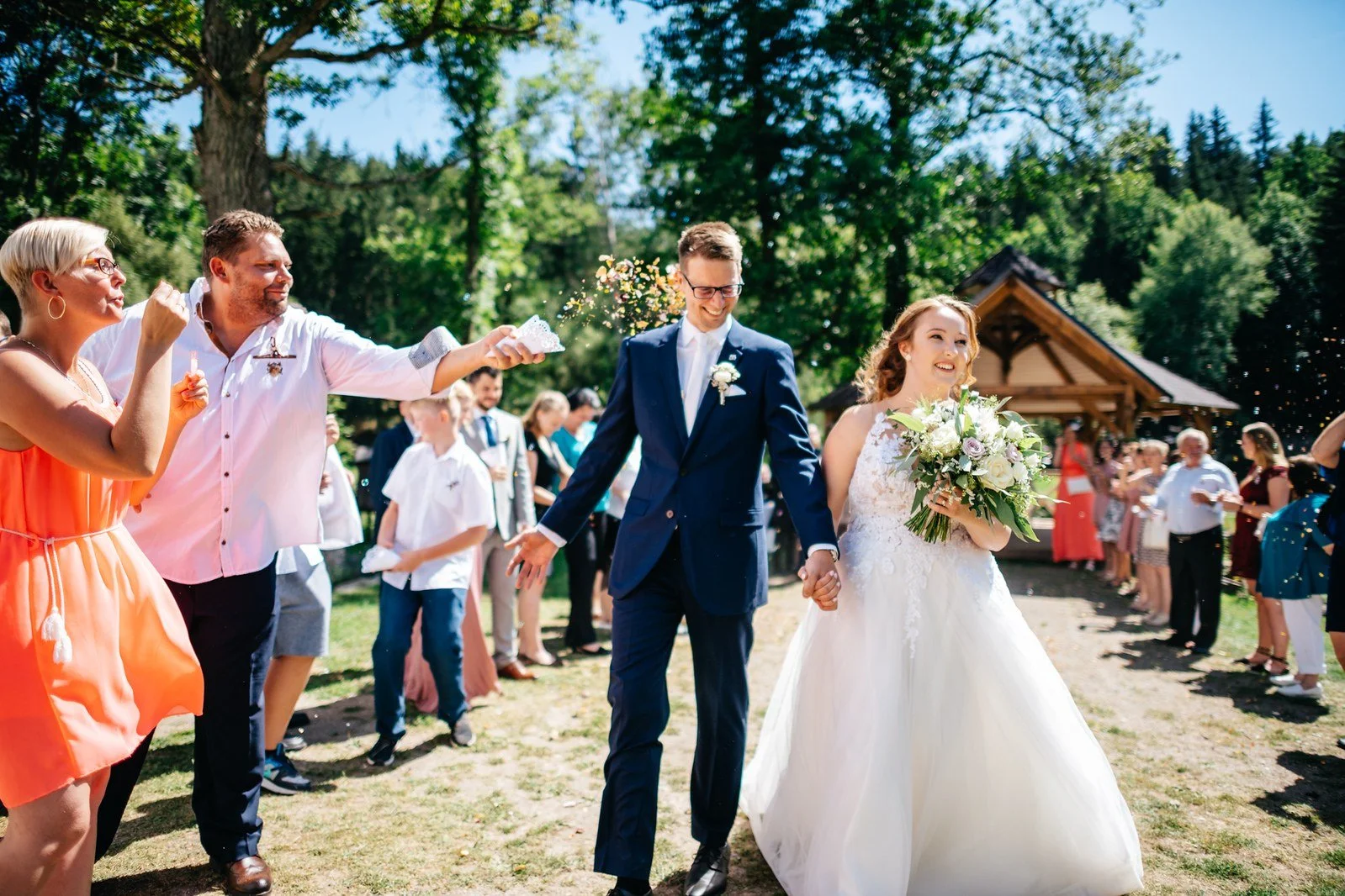Detail zachycuje ruce páru při navlékání prstenu během venkovního obřadu.